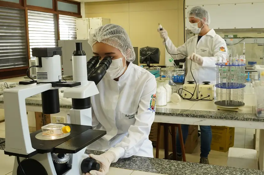 A imagem mostra um laboratório, uma pesquisadora realiza observações em um microscópio, utilizando equipamentos de proteção individual, enquanto outro profissional executa procedimentos técnicos ao fundo, em um ambiente equipado para análises científicas.