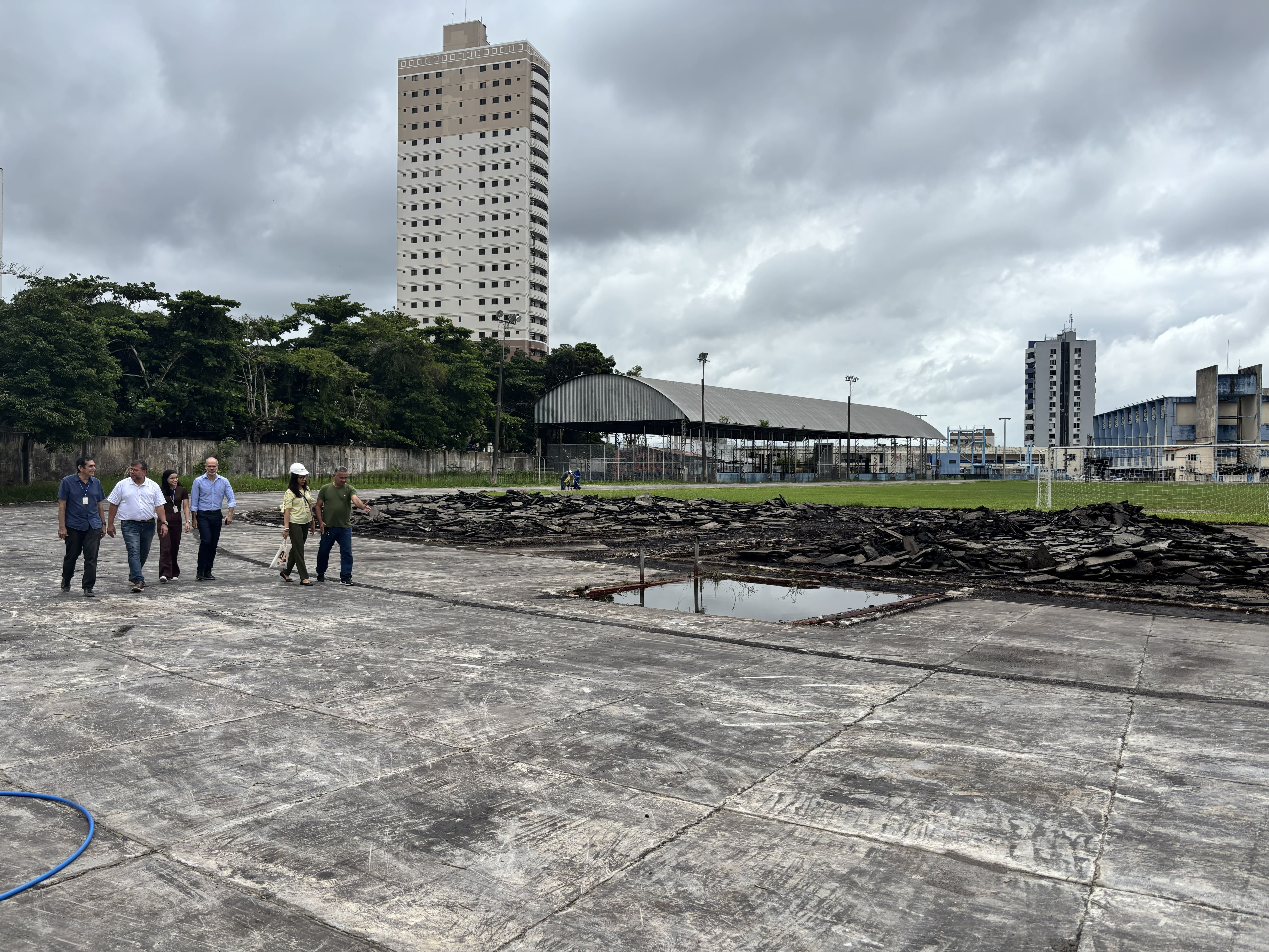 Um grupo de pessoas caminha por uma ampla área de concreto que aparenta estar em processo de reforma. Ao lado, há uma grande pilha de telhas removidas e um espaço com água acumulada. Ao fundo, observa-se um campo com traves de futebol, uma quadra coberta e prédios altos, sob um céu nublado.