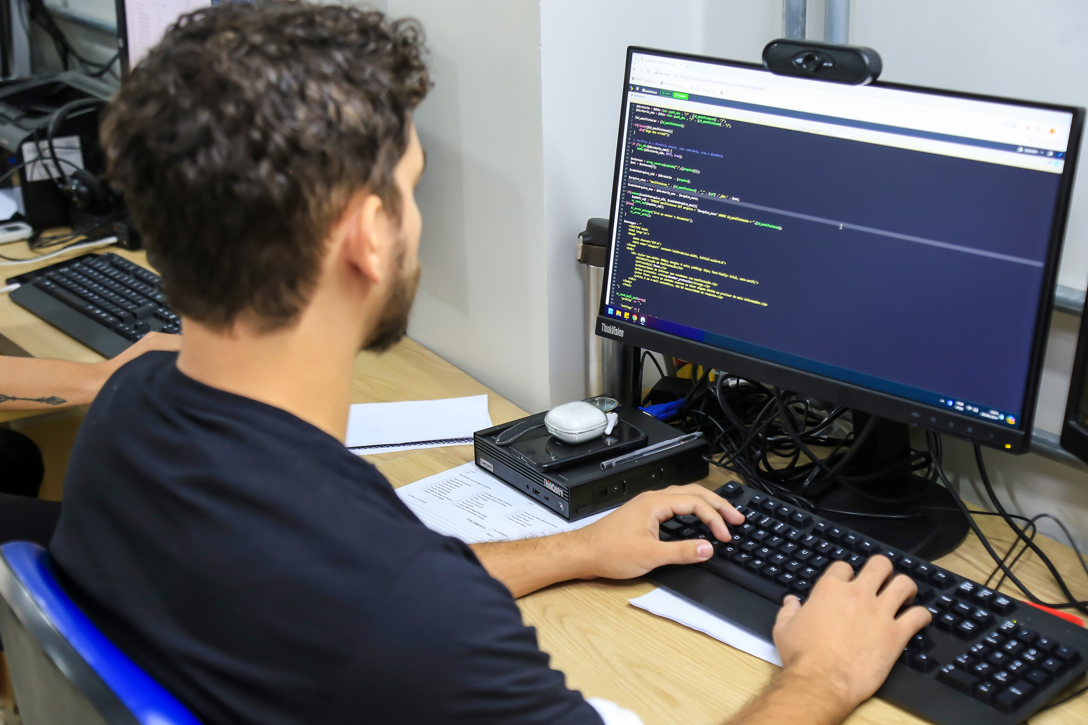 A imagem mostra um homem sentado em frente a um computador, trabalhando com programação. Na tela do monitor, há um editor de código com linhas escritas em linguagem de programação, provavelmente HTML, CSS ou JavaScript. Ele está em um ambiente de escritório ou sala de aula, sugerindo uma atividade de desenvolvimento de software ou aula prática de engenharia de software.