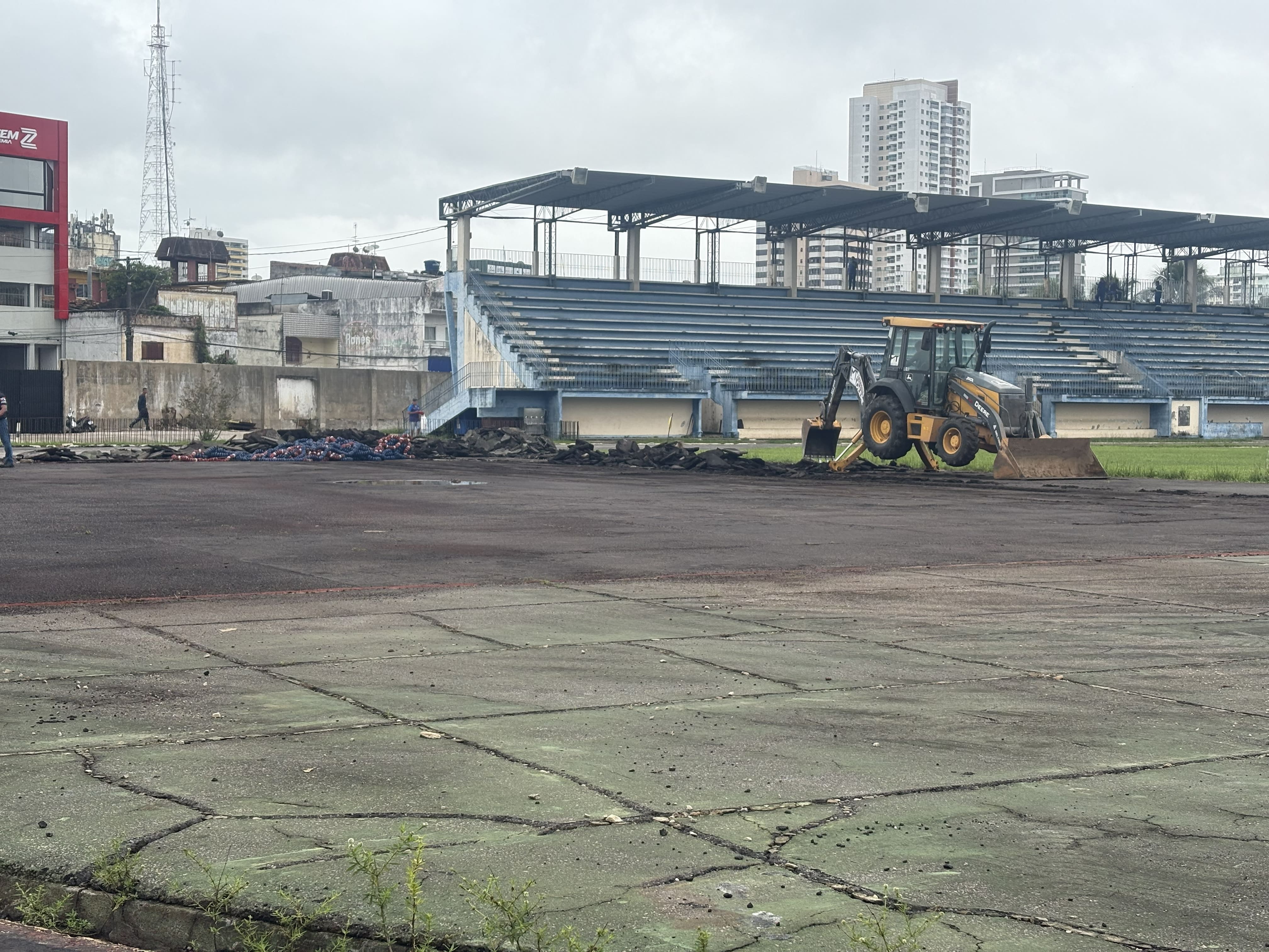 Uma retroescavadeira está parada sobre uma área esportiva em processo de reforma, ao lado de pilhas de material removido do piso. Ao fundo, há arquibancadas cobertas de um estádio ou quadra esportiva, além de prédios urbanos e uma torre de comunicação. O céu está nublado e o local apresenta sinais de desgaste no pavimento.