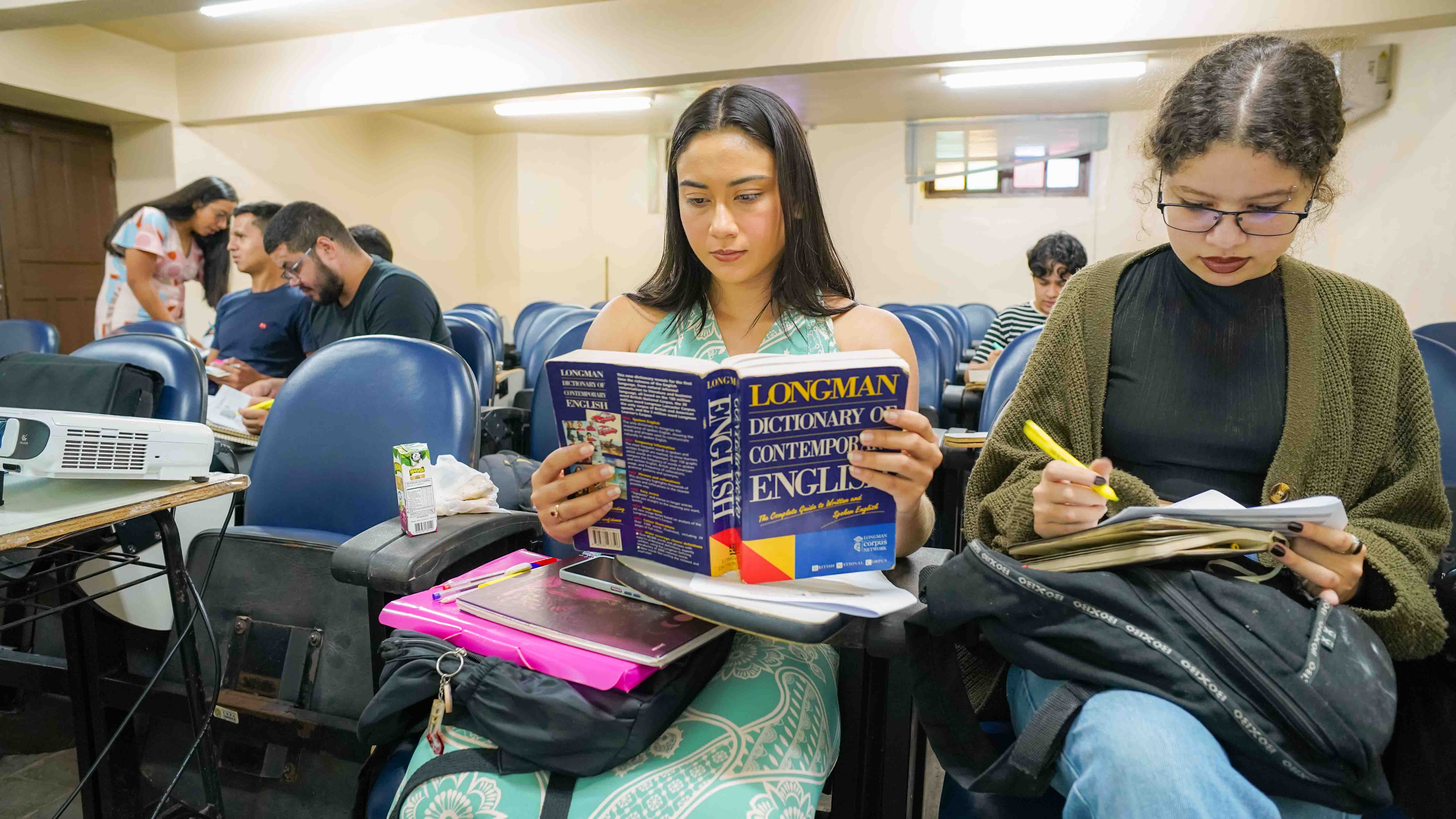 Estudantes sentados em uma sala de aula, com destaque para uma jovem ao centro lendo atentamente um exemplar do dicionário de inglês