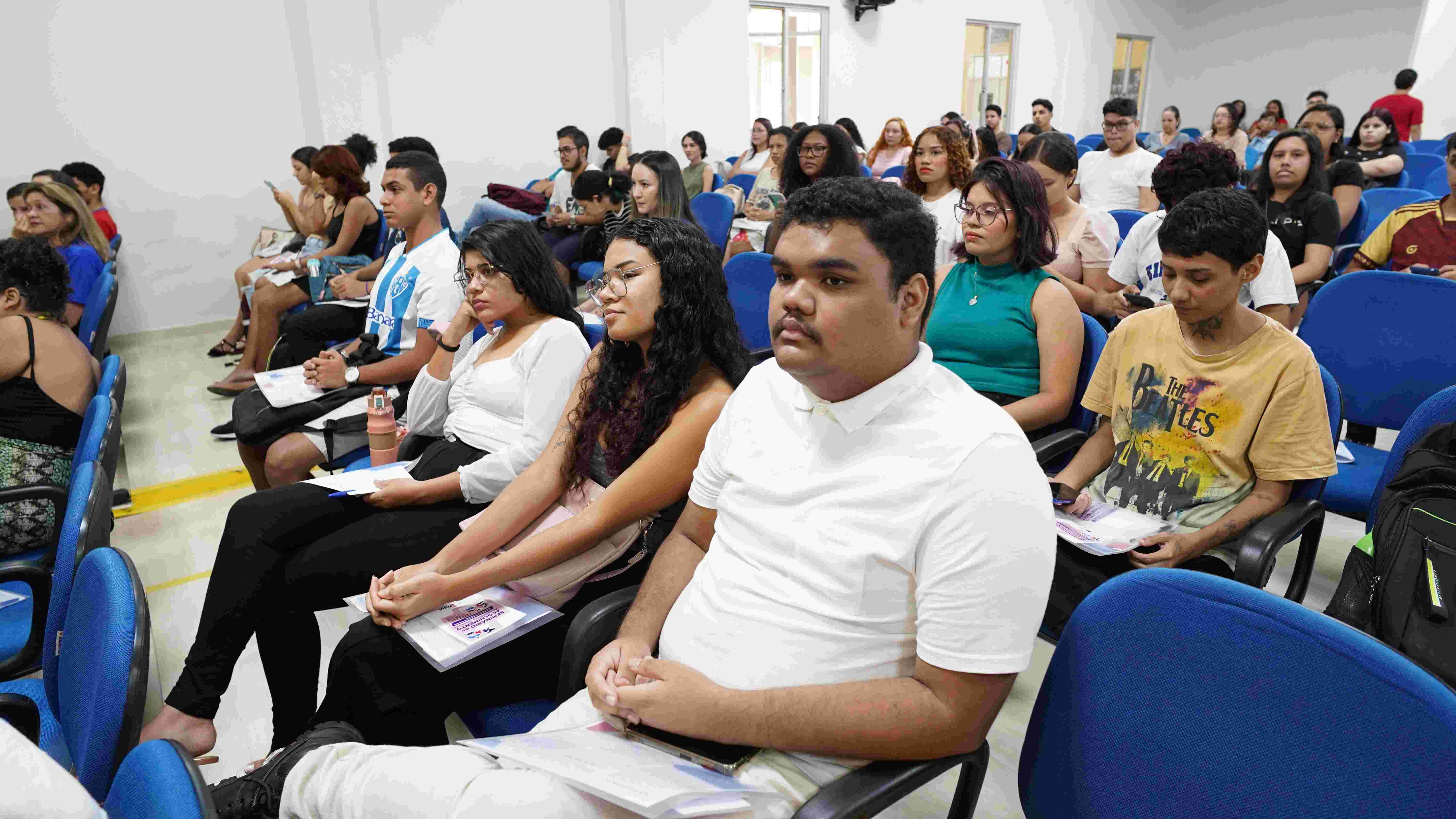 Fotografia mostra estudantes sentados em um auditório com cadeiras azuis durante uma atividade acadêmica. Em primeiro plano, jovens acompanham atentamente a programação enquanto seguram materiais impressos. Ao fundo, outros participantes ocupam diferentes fileiras do espaço, em ambiente iluminado por luz branca e paredes claras.