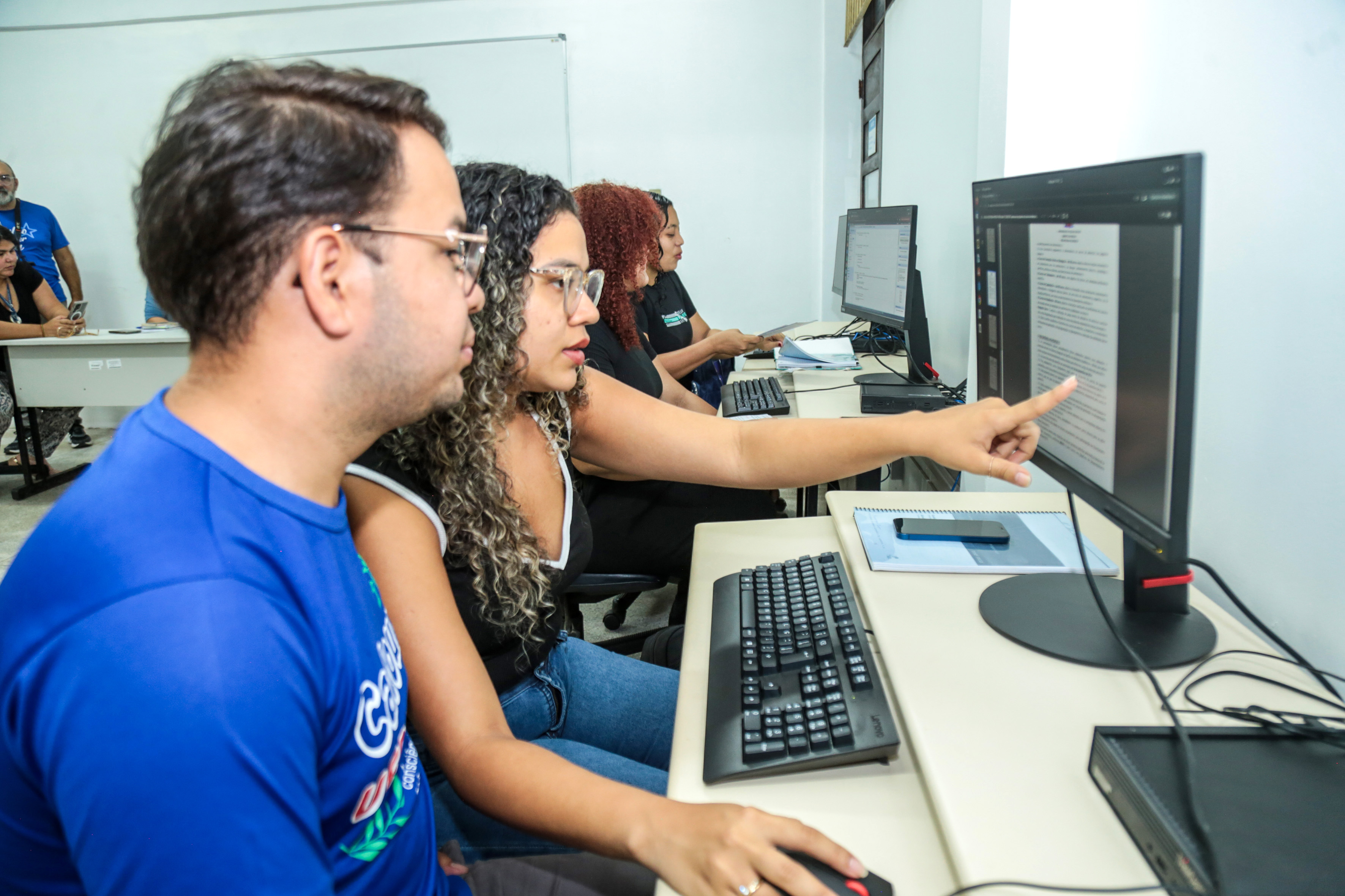 A imagem mostra dois jovens estudantes em uma sala, olhando para a tela de um computador, que exibe um documento. Um dos jovens aponta o dedo para a tela.
