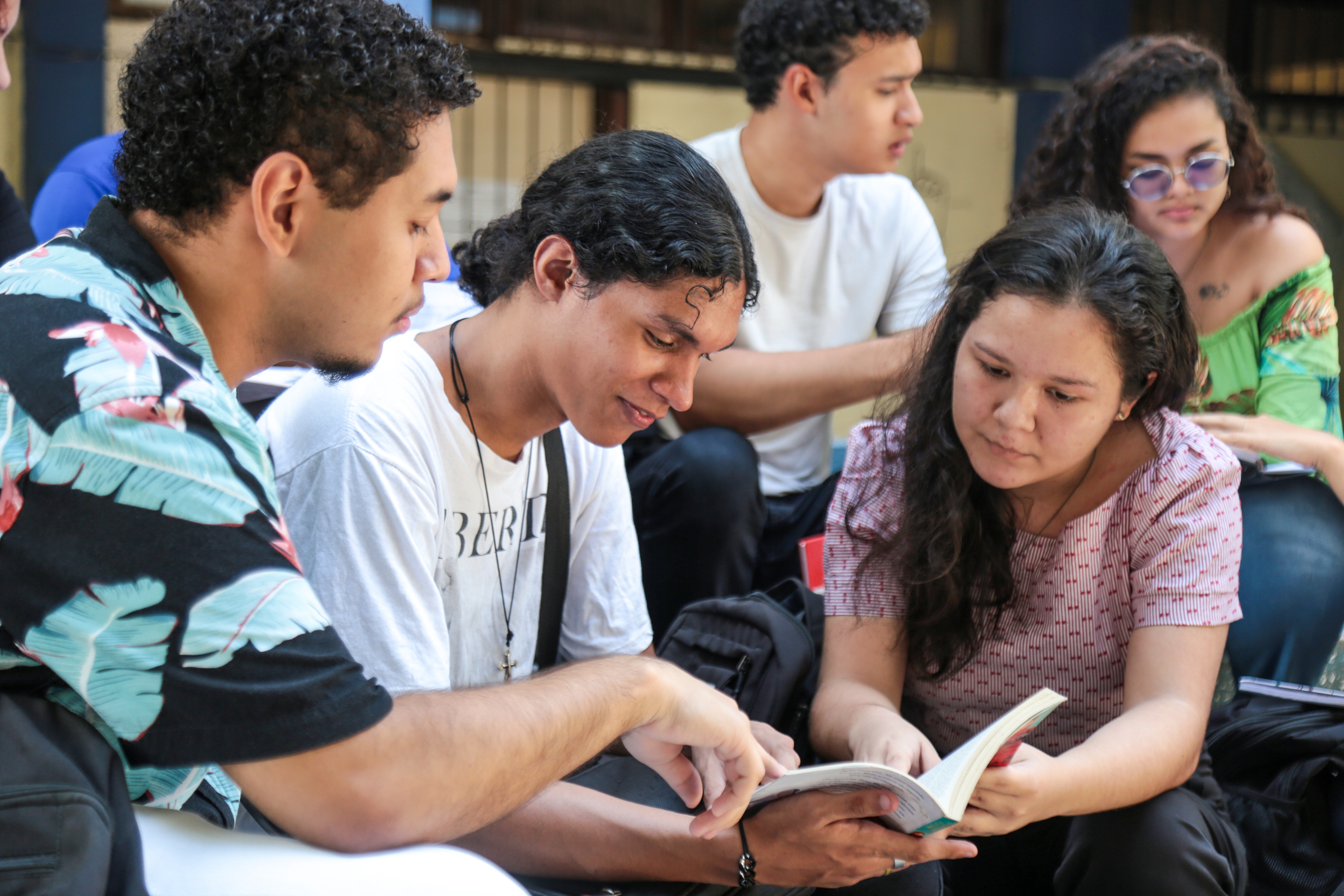 A imagem mostra um grupo de estudantes sentados próximos uns aos outros, envolvidos em uma sessão de estudos. À esquerda, uma pessoa está usando uma camisa com estampa floral e aponta para um livro. Ao lado dessa pessoa, há outra vestindo uma camiseta branca, olhando para baixo. No primeiro plano, uma mulher com cabelo longo está usando uma camisa rosa e também está olhando para papéis ou um livro em suas mãos. Ao fundo, outras pessoas podem ser vistas conversando.