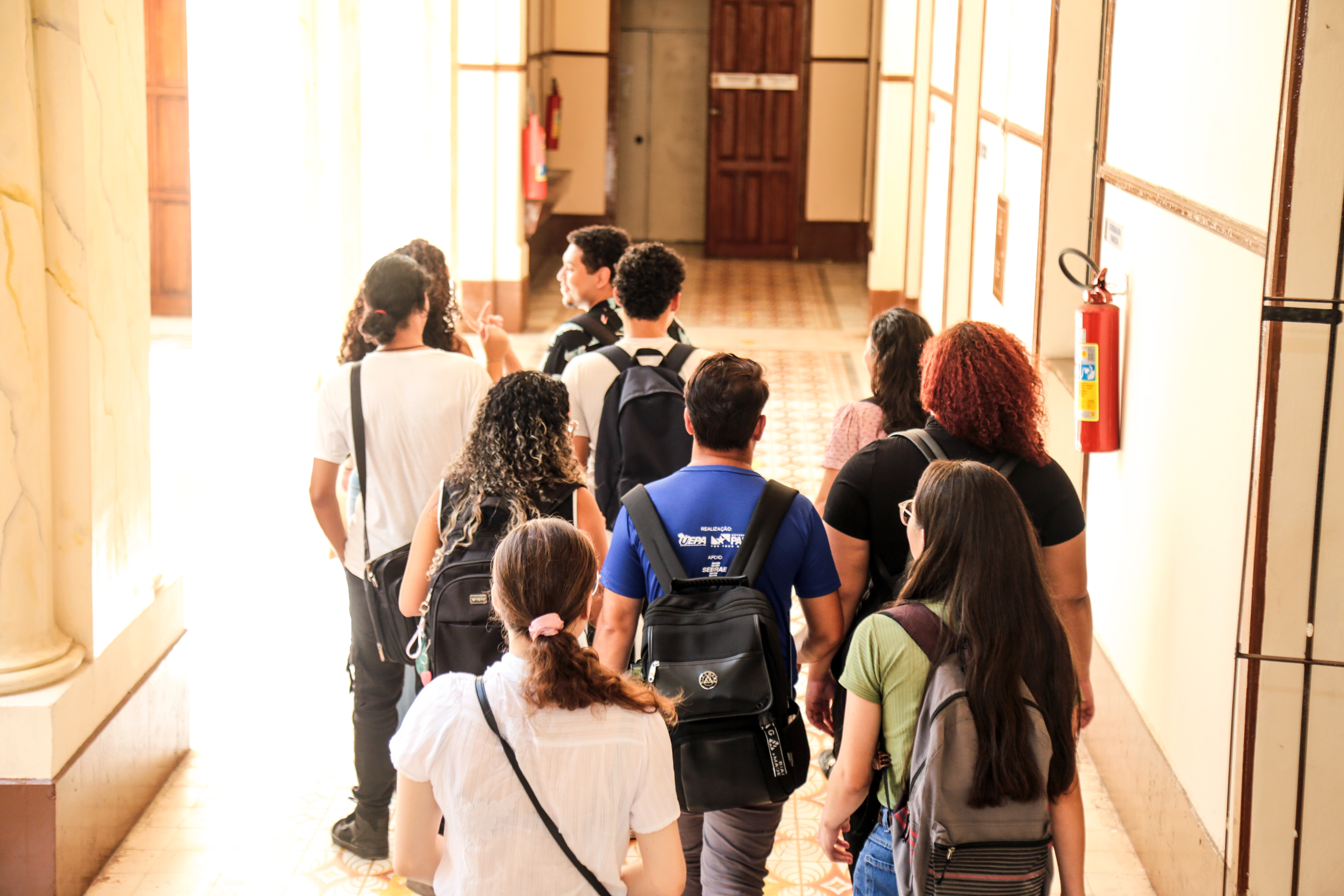 A imagem mostra um grupo de estudantes caminhando por um corredor iluminado e amplo. Eles estão de costas para a câmera e carregam mochilas, que indica um ambiente acadêmico. O corredor possui piso com detalhes geométricos e paredes claras com acabamentos de madeira. Há portas de madeira ao fundo e uma luz natural que entra pelas grandes  aberturas laterais.