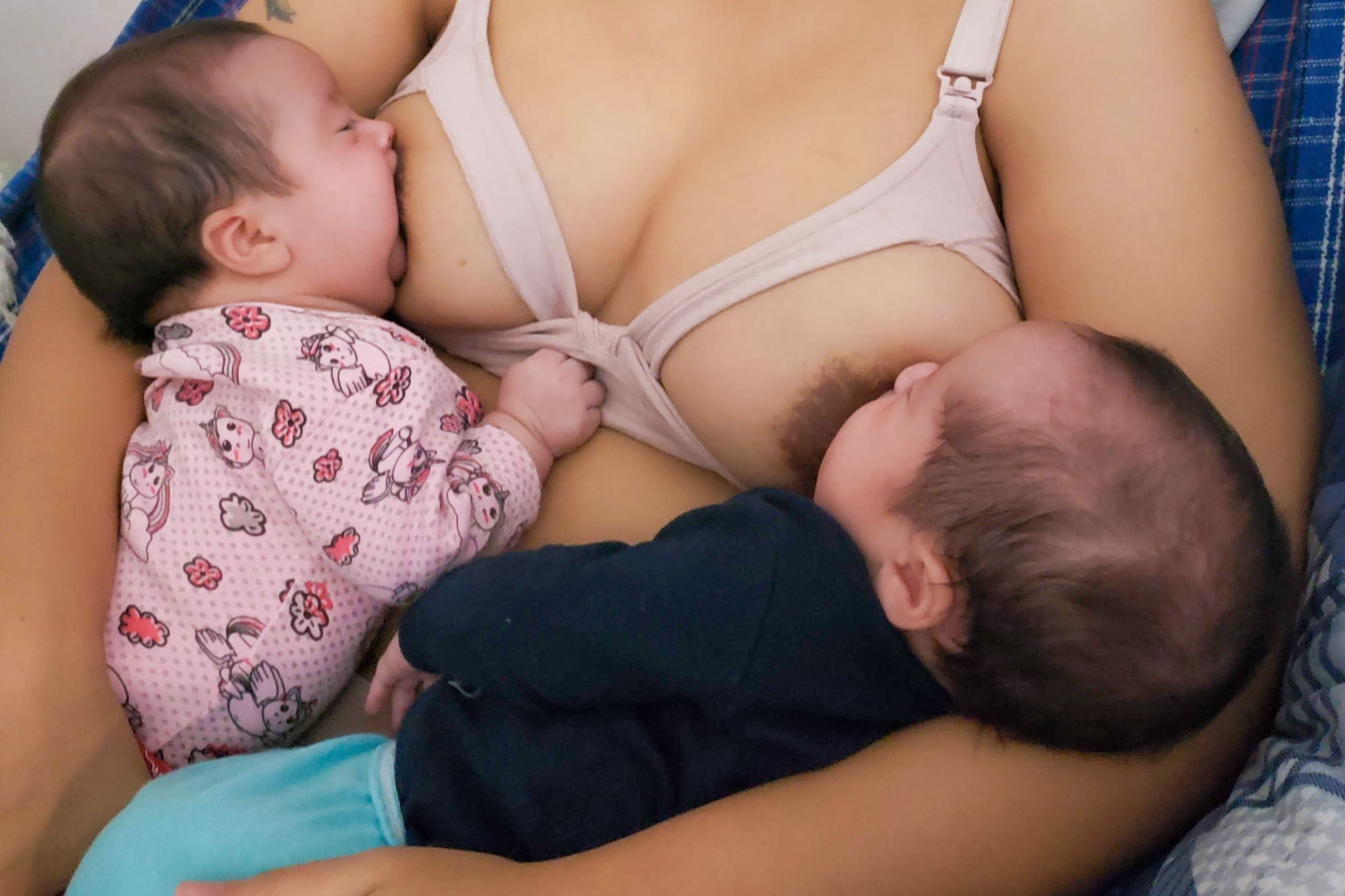 A imagem mostra uma mulher amamentando dois bebês ao mesmo tempo, cada um posicionado em um dos seios. Um dos bebês veste roupa rosa estampada com unicórnios e flores, enquanto o outro usa blusa azul-escura e calça azul-clara. A cena transmite cuidado, vínculo e afeto materno.