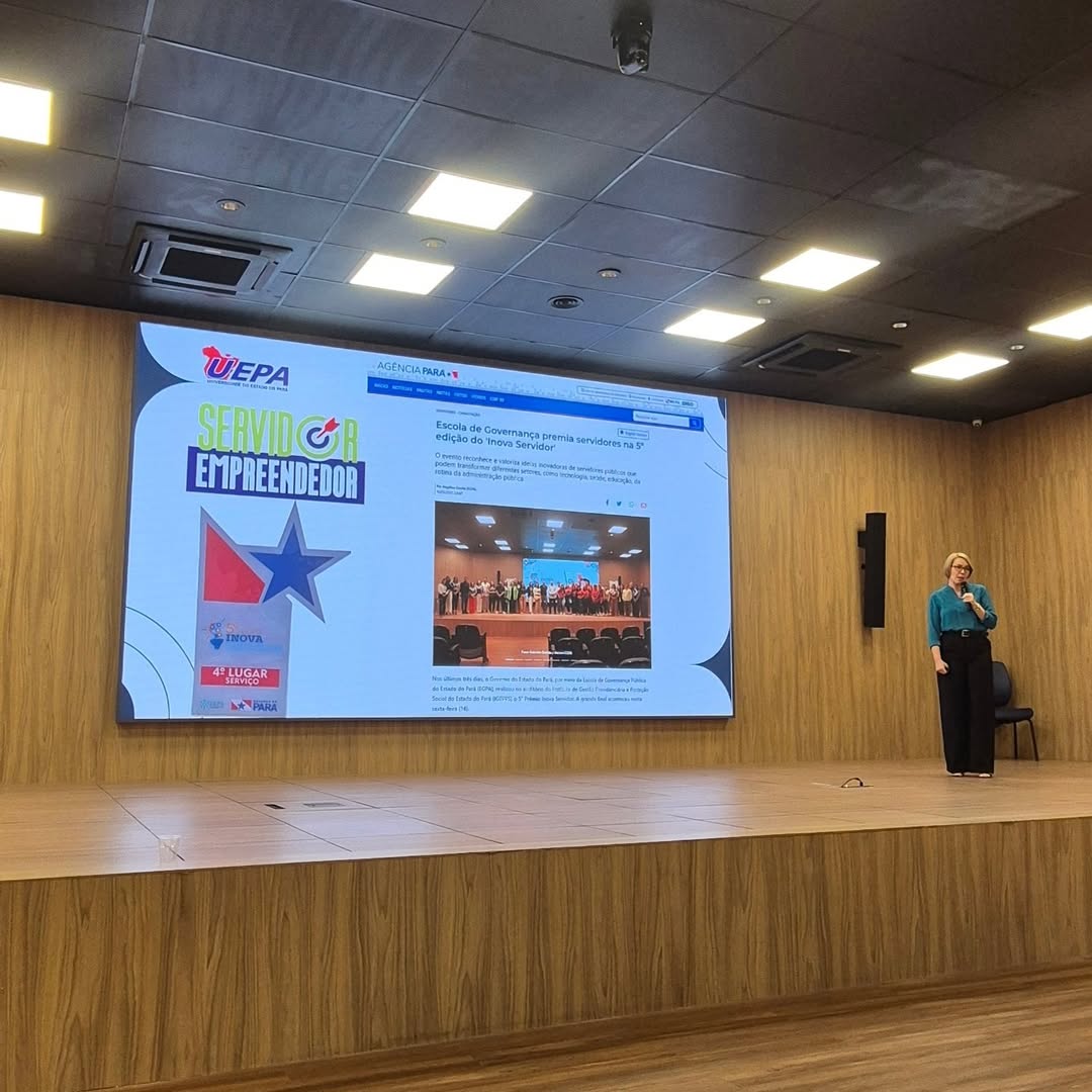 A imagem apresenta um registro desfocado e parcialmente obstruído, em que cabelos cobrem grande parte da lente. Ao fundo, é possível identificar um ambiente interno iluminado, com painel institucional exibindo a marca da Uepa e um espaço que parece ser de apresentação ou evento, com elementos visuais e materiais expostos.