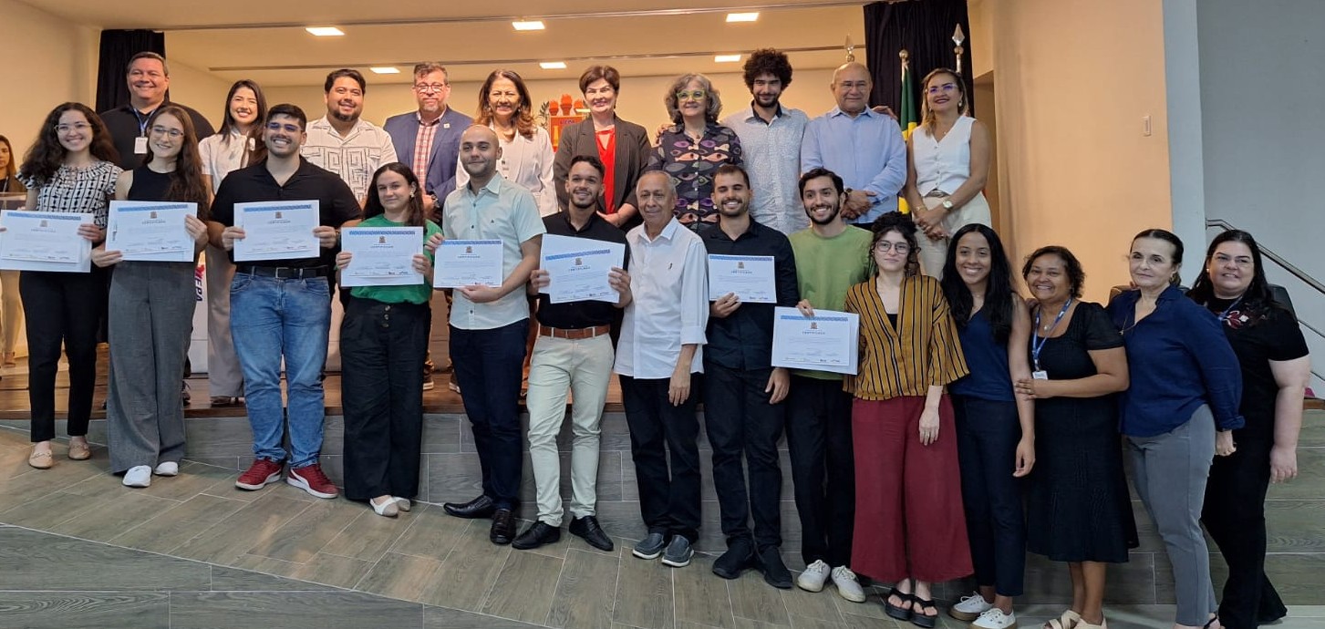 A imagem mostra um grupo de pessoas reunidas em um auditório, em clima de celebração. Na frente, participantes exibem certificados, enquanto atrás deles estão docentes e gestores, todos sorridentes, em um registro oficial do evento.