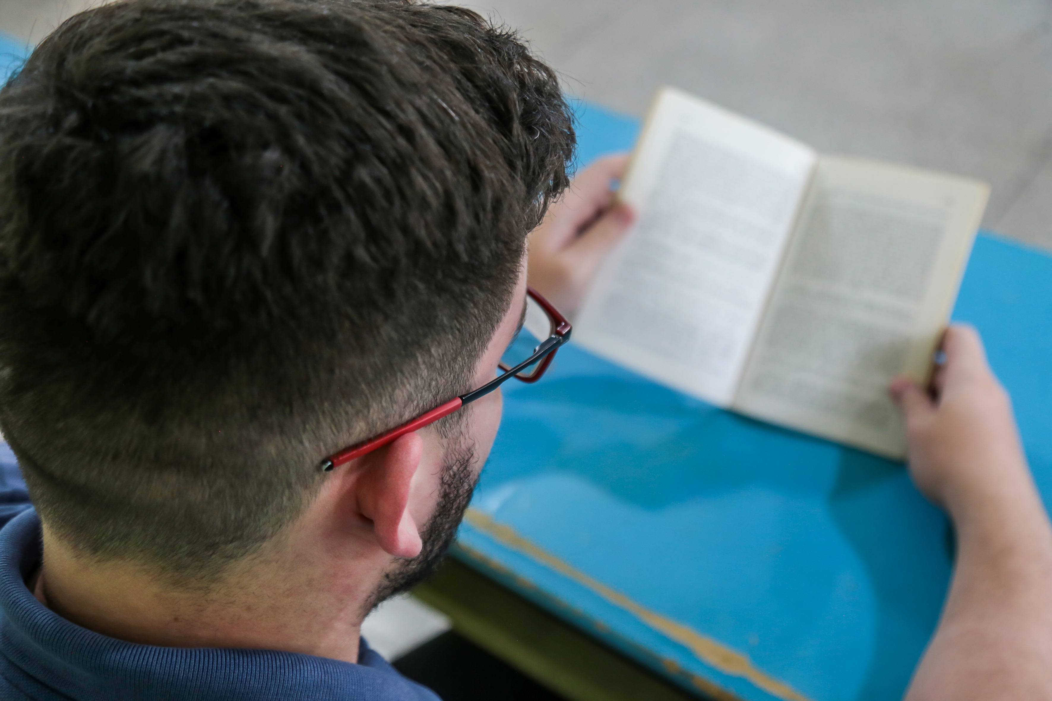 A imagem mostra uma pessoa vista por trás, sentada e lendo um livro aberto sobre uma mesa azul. Ela usa óculos de armação vermelha e veste uma camisa azul.