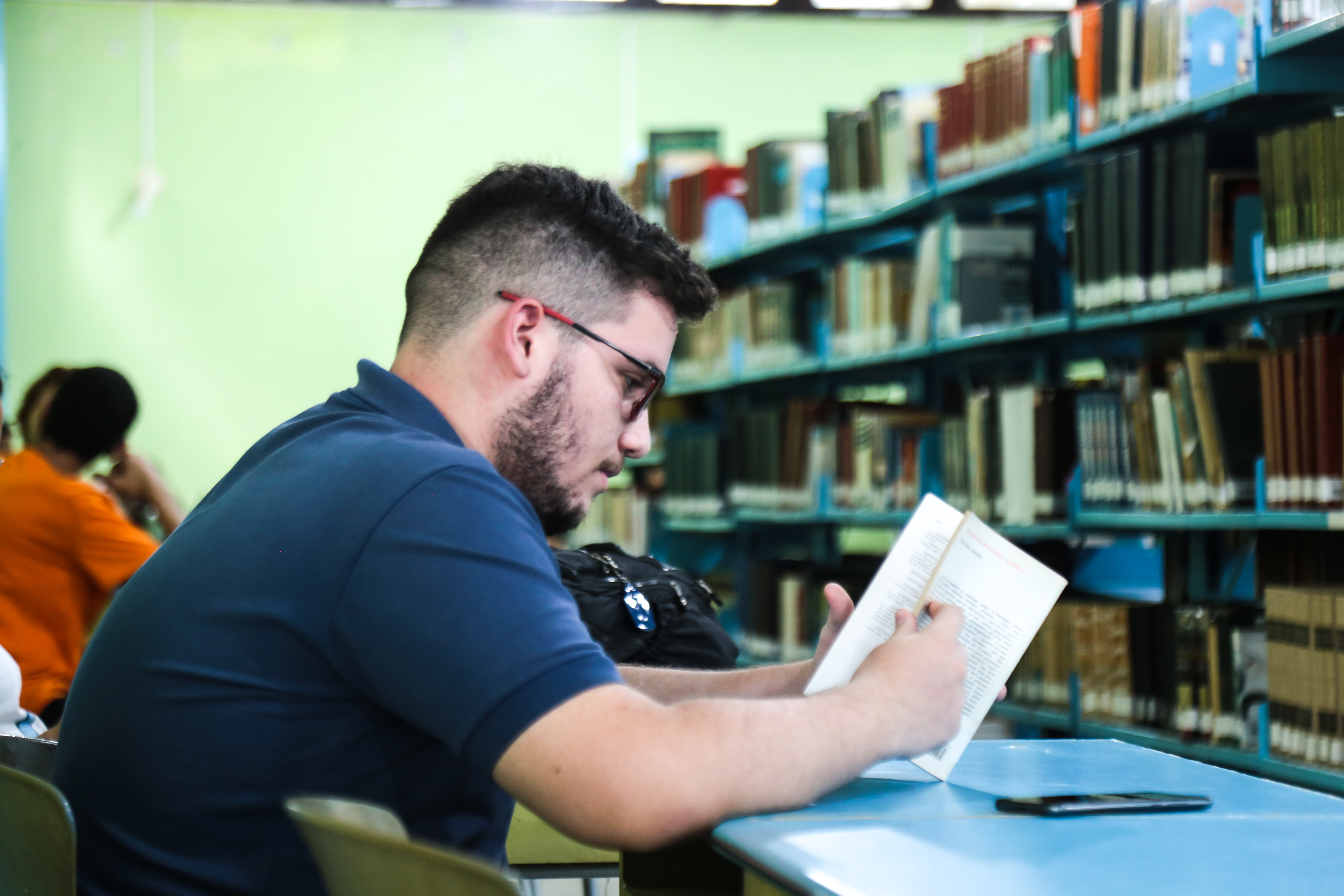 Um estudante está sentado em uma mesa de biblioteca, concentrado na leitura de um livro, com estantes cheias de obras ao fundo.