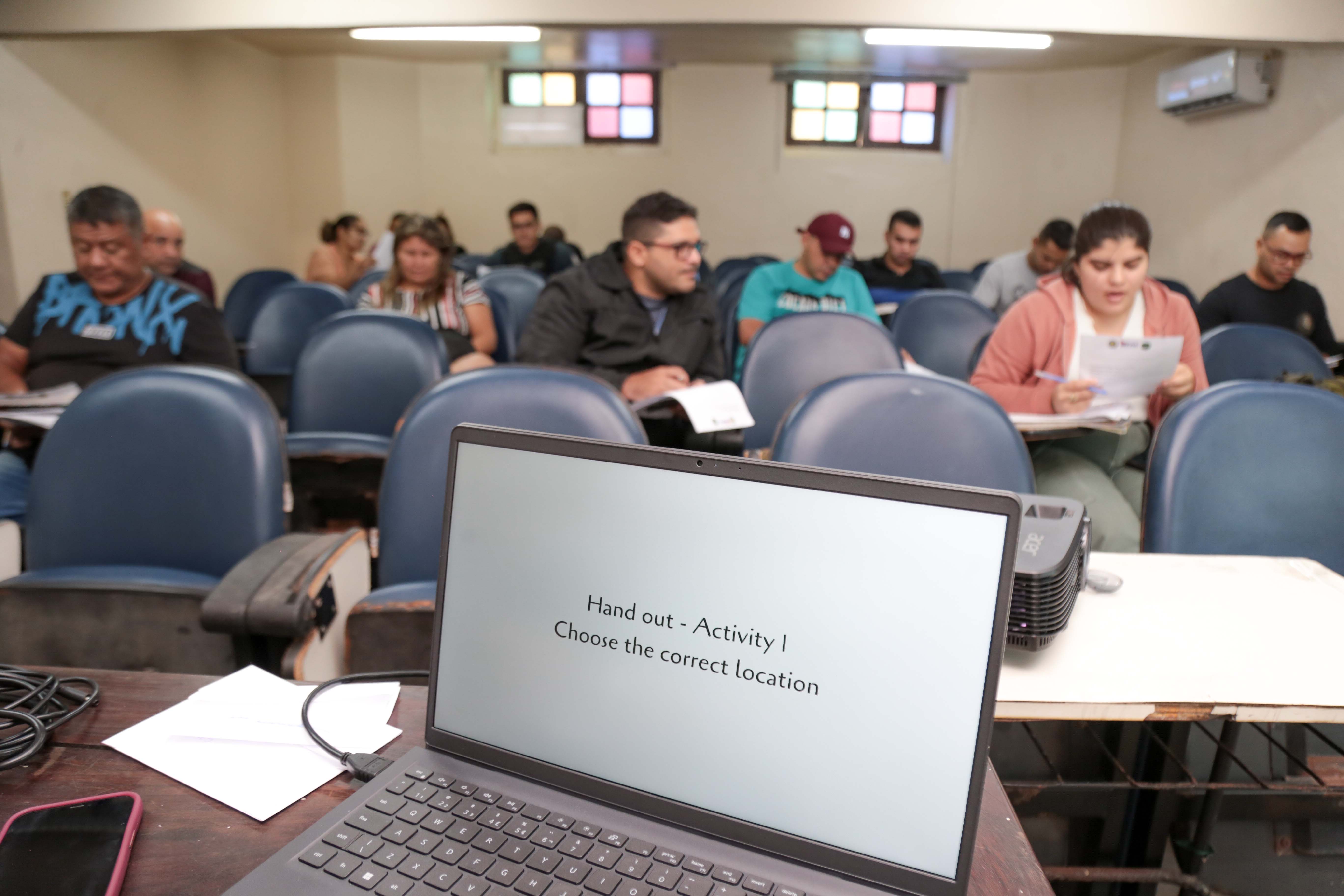 A imagem mostra uma sala de aula com alunos sentados em carteiras azuis, concentrados em uma atividade impressa. Em primeiro plano, um notebook exibe a mensagem "Hand out – Activity I – Choose the correct location". A sala tem iluminação fluorescente, janelas com vidros coloridos e um ar-condicionado na parede.