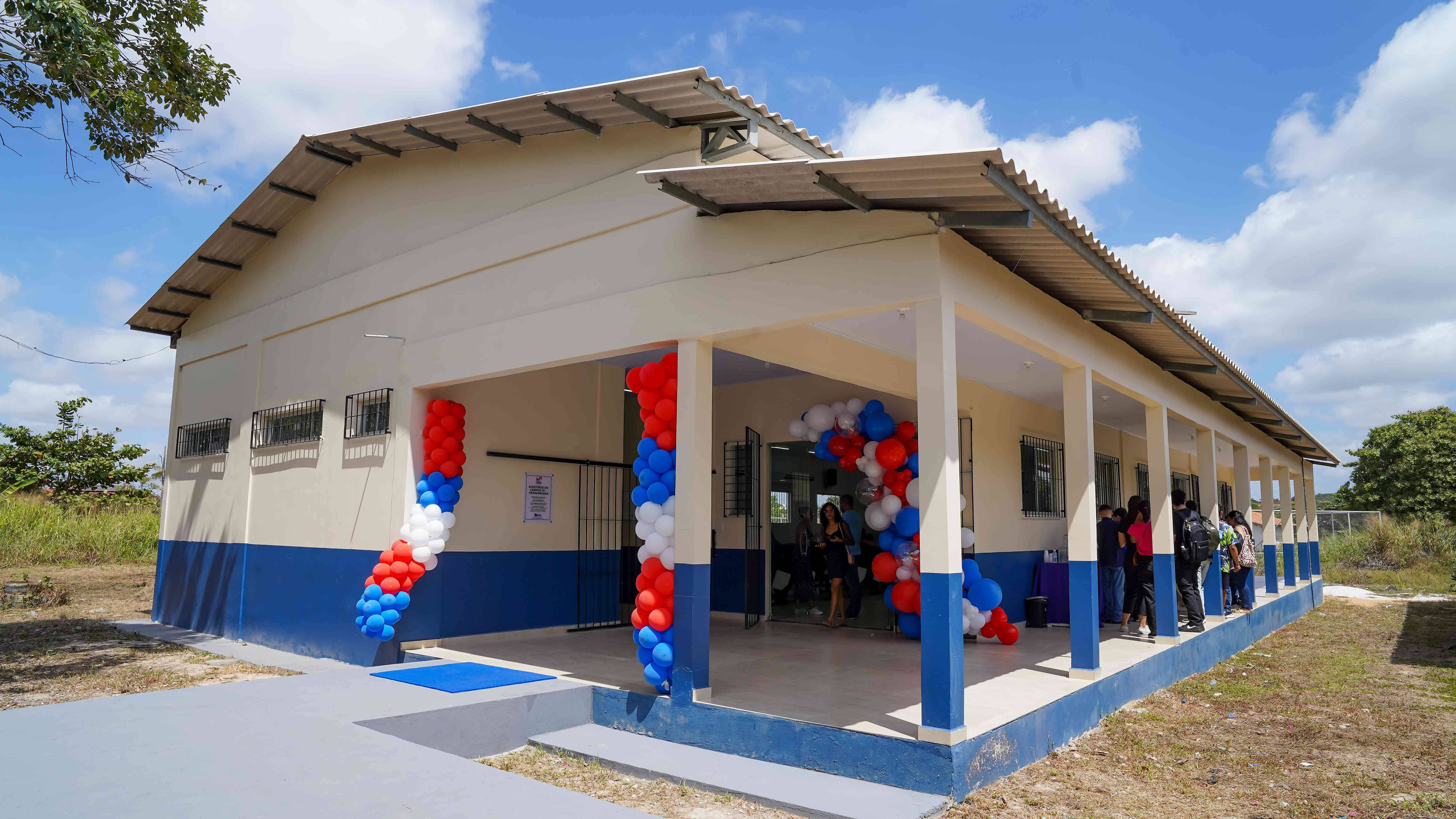 A imagem mostra um prédio de um único andar com fachada branca e detalhes em azul ao redor das janelas e portas.  Há decorações com balões vermelhos, brancos e azuis dispostos em grupos ao redor da entrada, para o evento de inauguração. O prédio está situado em uma área gramada com poucas árvores ao fundo, sob um céu parcialmente nublado.