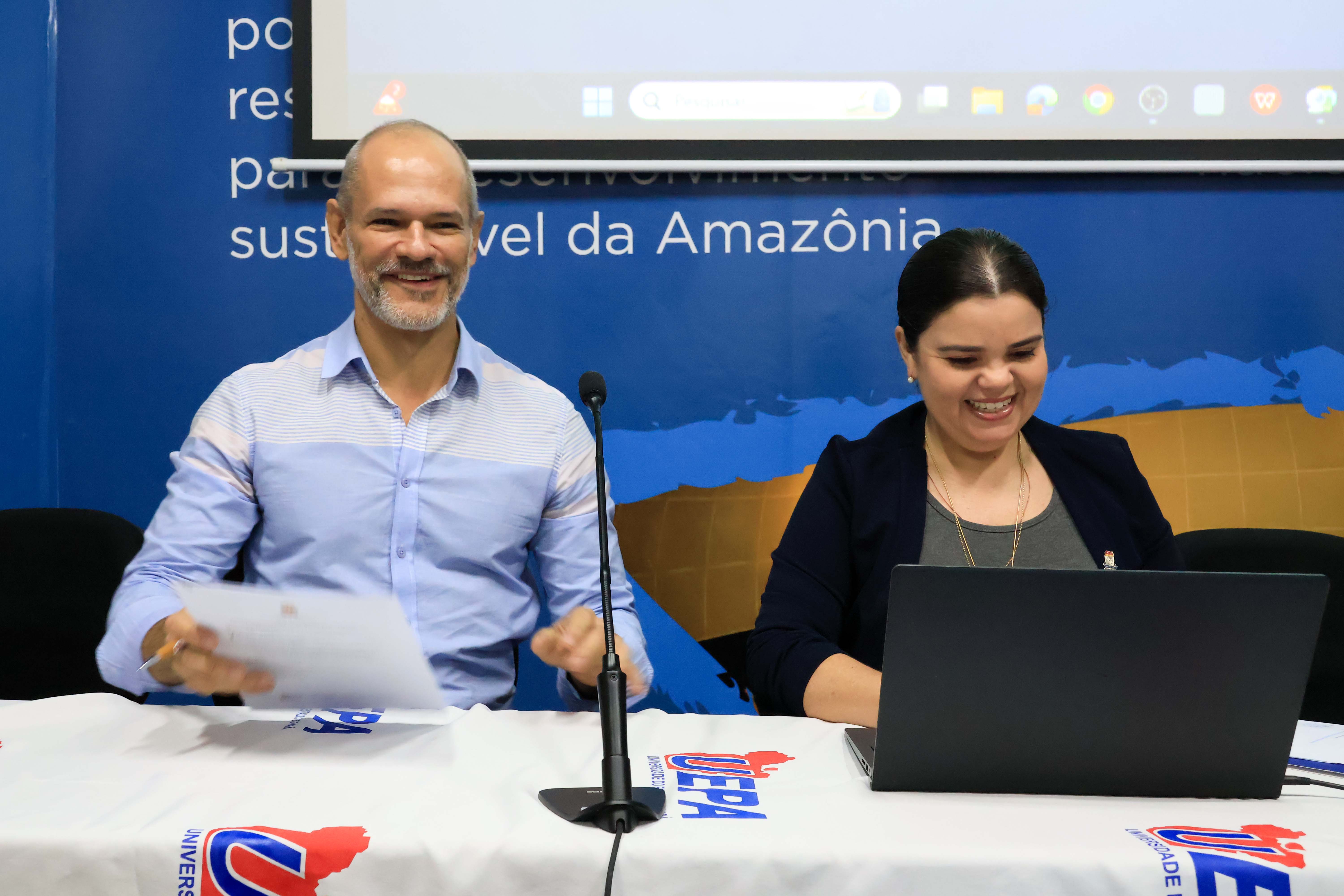 Dois representantes das universidades participam sorridentes de uma reunião ou evento institucional. Sentados à mesa, com microfone e notebook à frente, eles demonstram envolvimento e bom humor. Ao fundo, há um painel azul com elementos visuais da UEPA e a frase “desenvolvimento sustentável da Amazônia”.