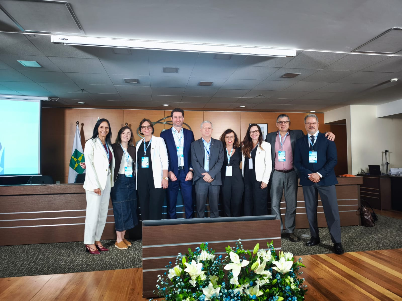 Grupo de nove pessoas posando lado a lado em um auditório ou sala de eventos institucionais, todas com crachás, sugerindo participação em um encontro ou programa acadêmico. Ao fundo, há uma mesa de conferência, bandeira institucional e tela de projeção; à frente, um arranjo de flores brancas. Os participantes vestem trajes formais e sorriem para a foto.