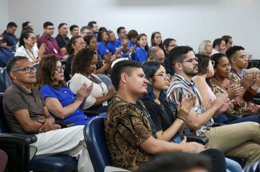 Uma plateia sentada em poltronas azuis de um auditório aplaude um evento. Em destaque na frente, um jovem de camisa marrom estampada, uma moça de cabelo azul e óculos, e um homem de camisa listrada. À esquerda, um homem de polo marrom e uma mulher de vestido azul-royal. Paredes brancas ao fundo.