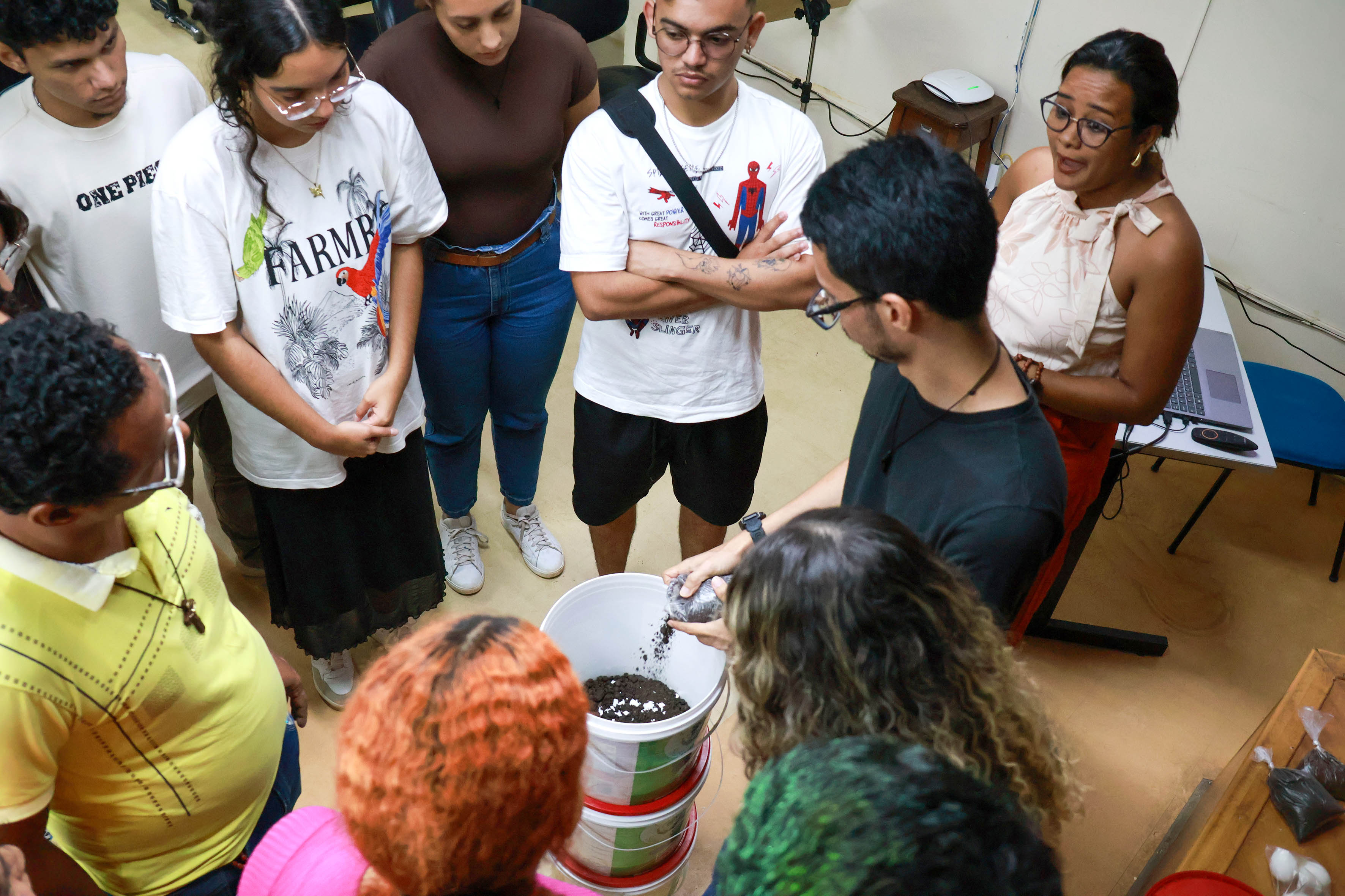 Na foto, um grupo de estudantes está reunido em torno de um balde com terra, acompanhando uma explicação prática sobre compostagem. Uma pessoa ao centro demonstra o processo, segurando um punhado de material orgânico, enquanto os demais observam com atenção. A cena se trata de uma atividade educativa e de conscientização ambiental.