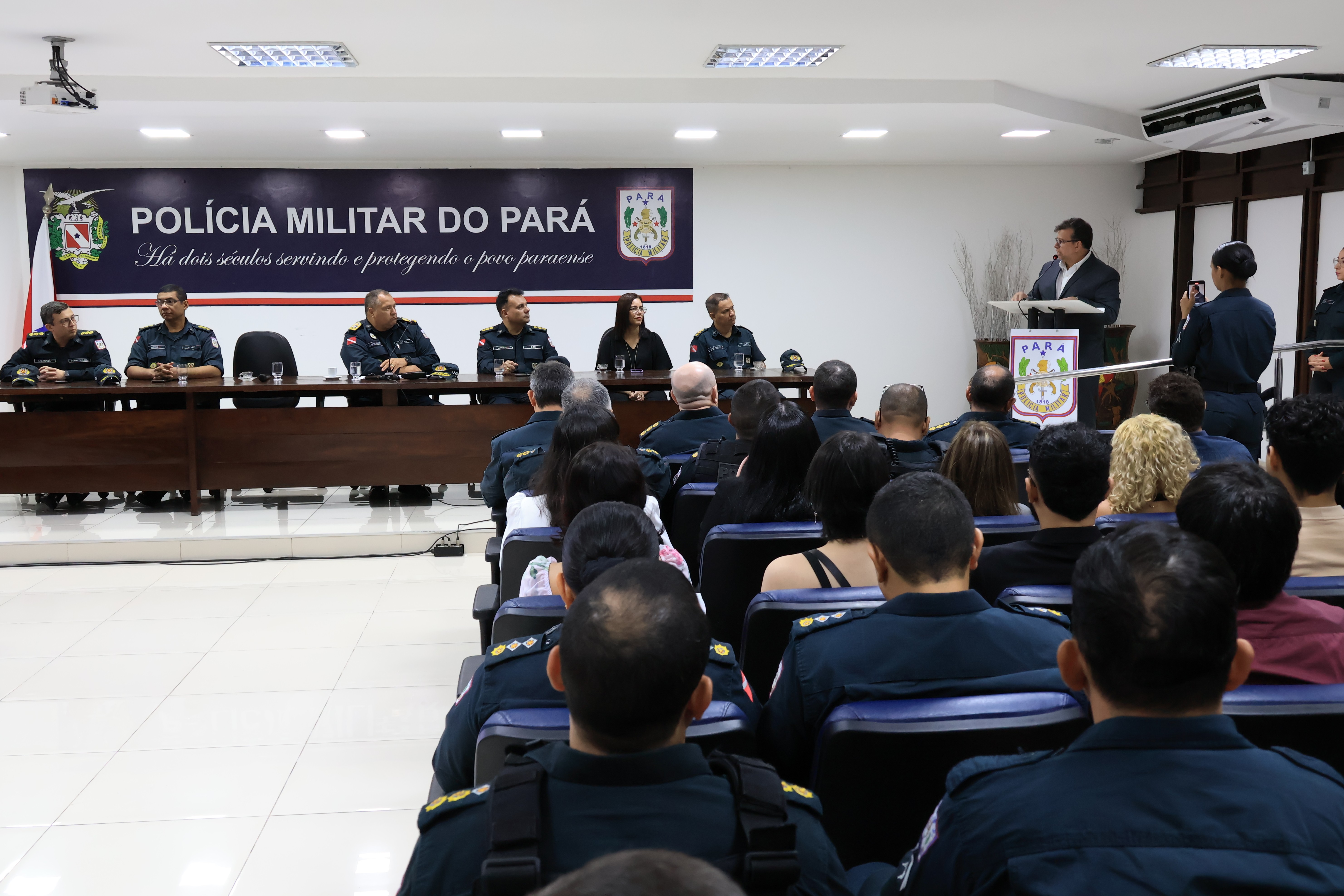 A imagem registra um momento de discurso durante uma cerimônia oficial da Polícia Militar do Pará. Um homem de terno fala ao púlpito, enquanto a mesa de autoridades acompanha atentamente. 