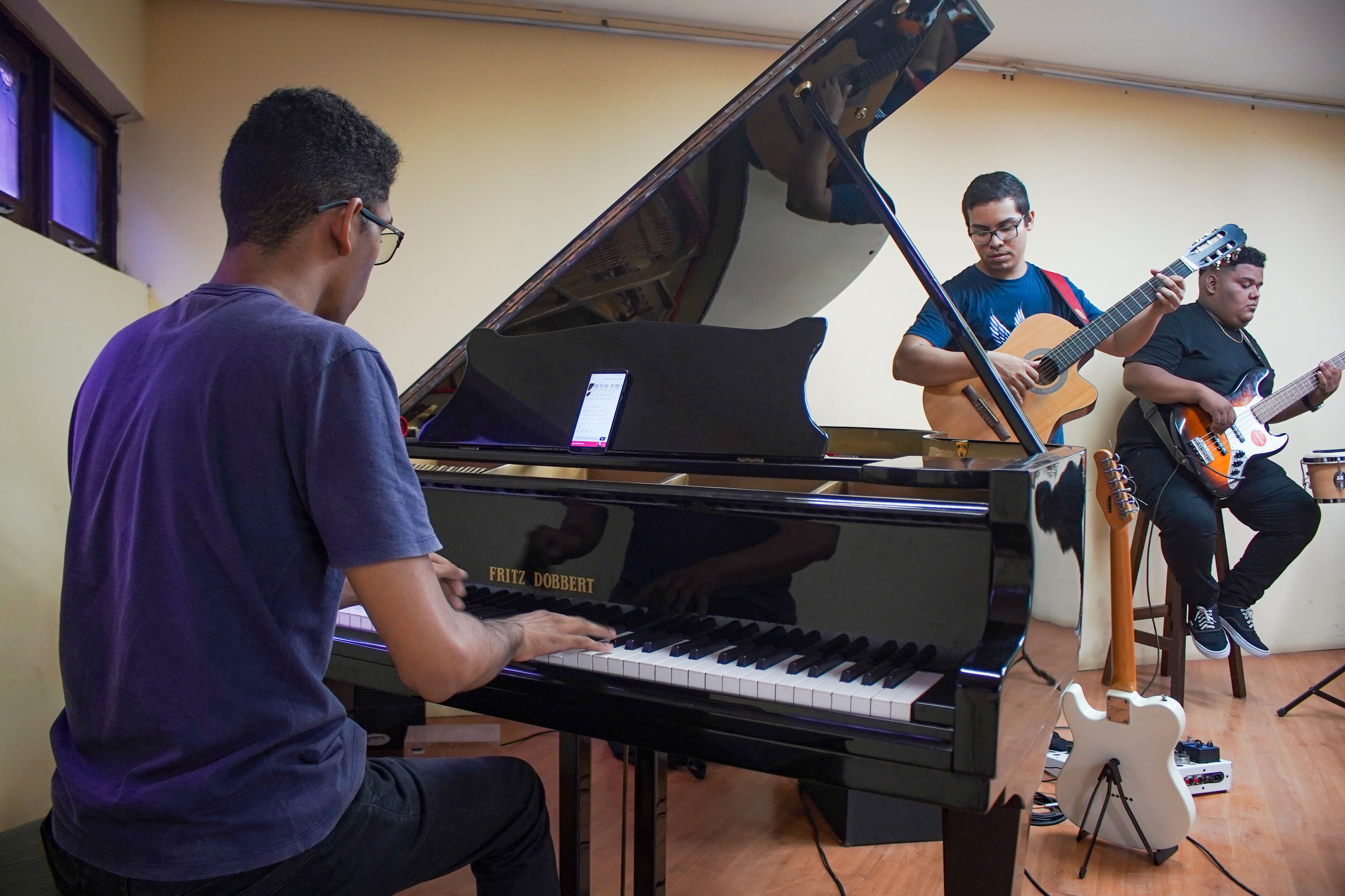 A imagem mostra três músicos em uma sala ensaiando: um toca piano de cauda (marca Fritz Dobbert), outro violão e o terceiro baixo elétrico. O pianista está de costas, enquanto os outros dois músicos estão sentados à frente, concentrados na execução.