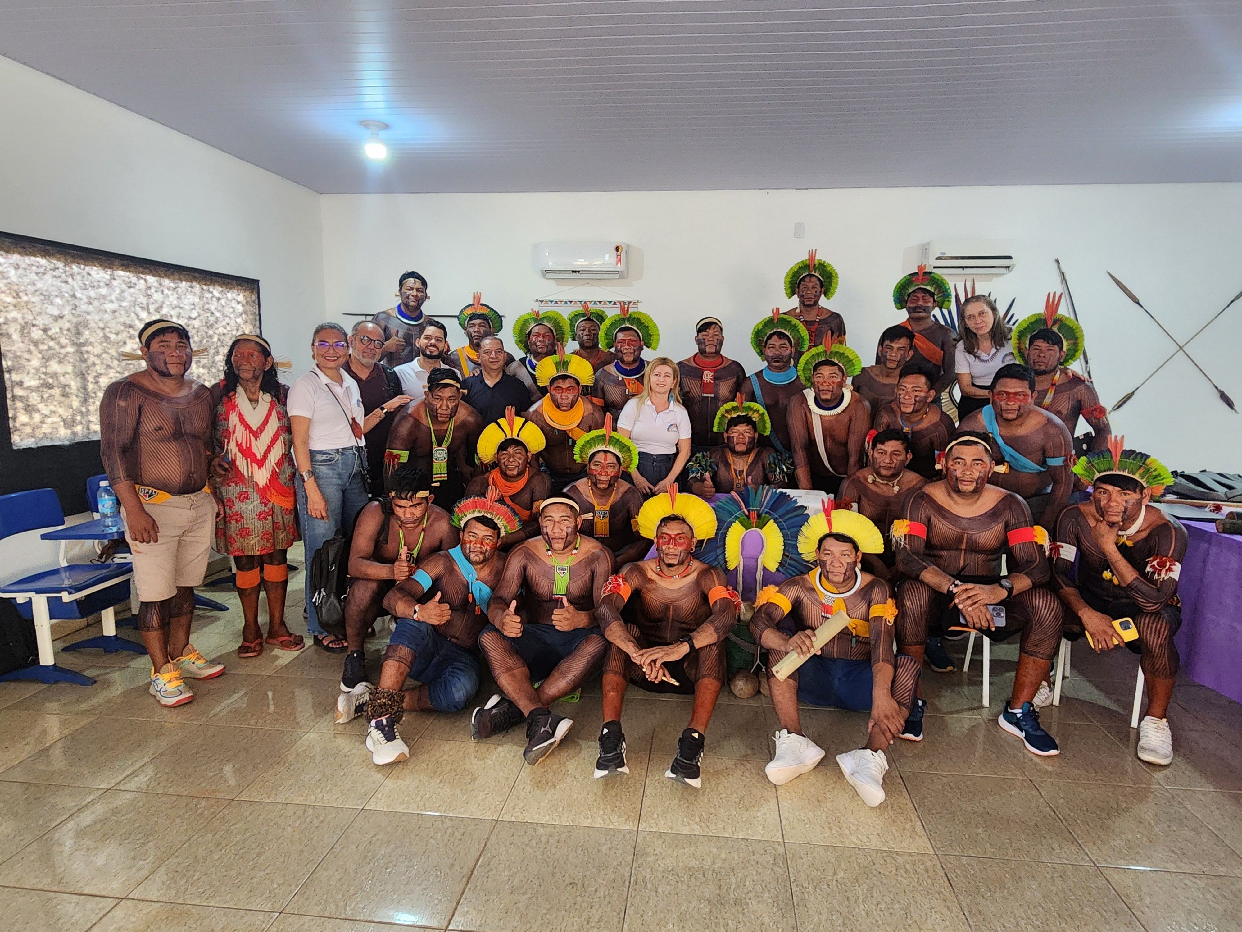 Na imagem, um grupo numeroso de pessoas posa reunido em um ambiente fechado, iluminado e organizado, com piso claro e paredes brancas. No centro da cena estão os acadêmicos Xikrin, vestidos com pinturas corporais tradicionais, cocares coloridos e adornos que evidenciam a cultura do povo. Eles estão acompanhados por professores, coordenadores e convidados, que aparecem ao fundo e nas laterais, compondo uma foto coletiva marcada pela celebração. Ao fundo, há ar-condicionados instalados e, na parede, enfeites 
