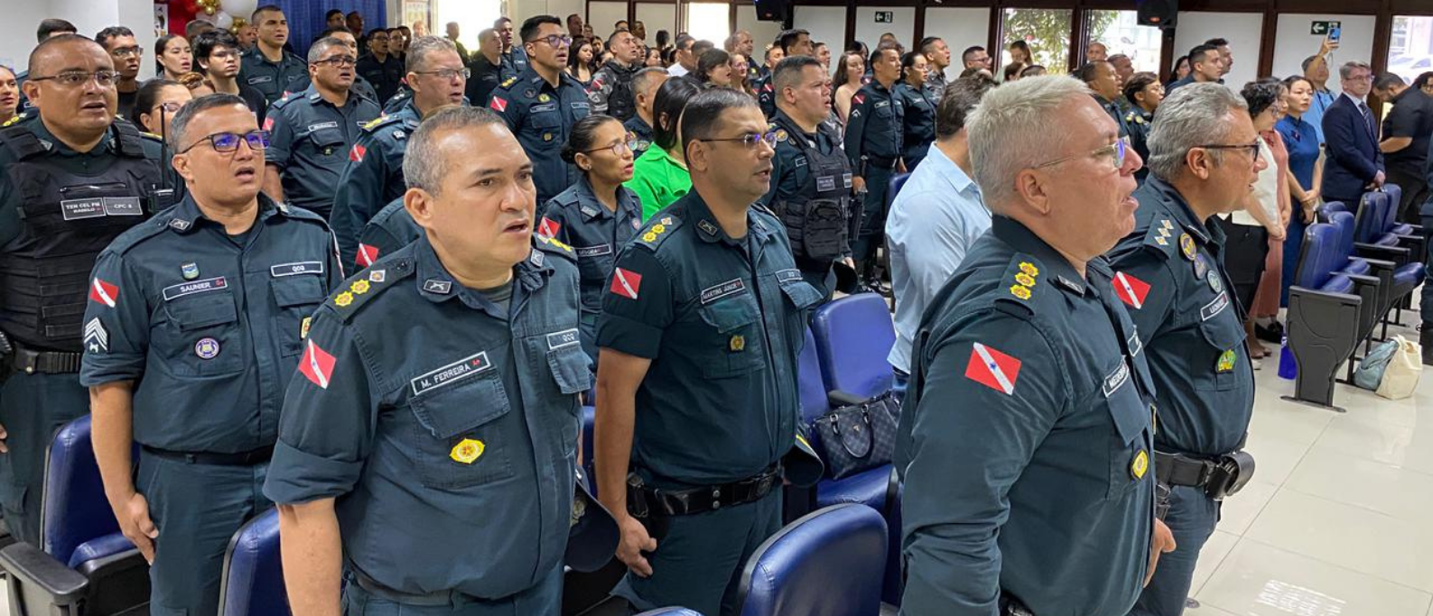 Policiais militares e convidados participam do momento solene da cerimônia, em pé, durante a execução do hino. O auditório está lotado e decorado com balões nas cores azul, vermelho e branco.