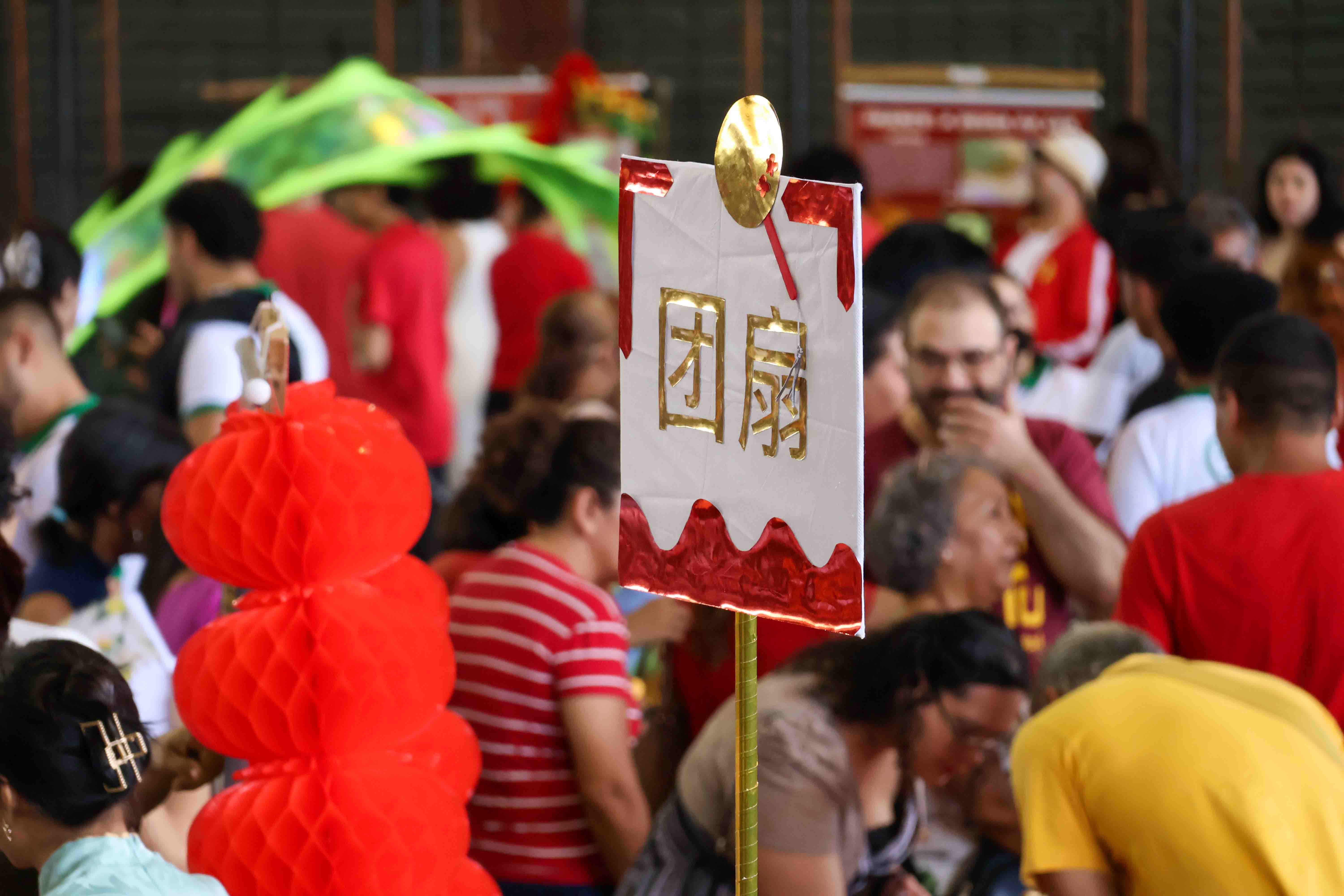 A foto mostra um espaço cheio de pessoas reunidas em um evento cultural. Em destaque, no centro, há uma placa branca e vermelha com caracteres chineses dourados. Ao lado, vê-se uma decoração em papel vermelho no formato de lanternas.