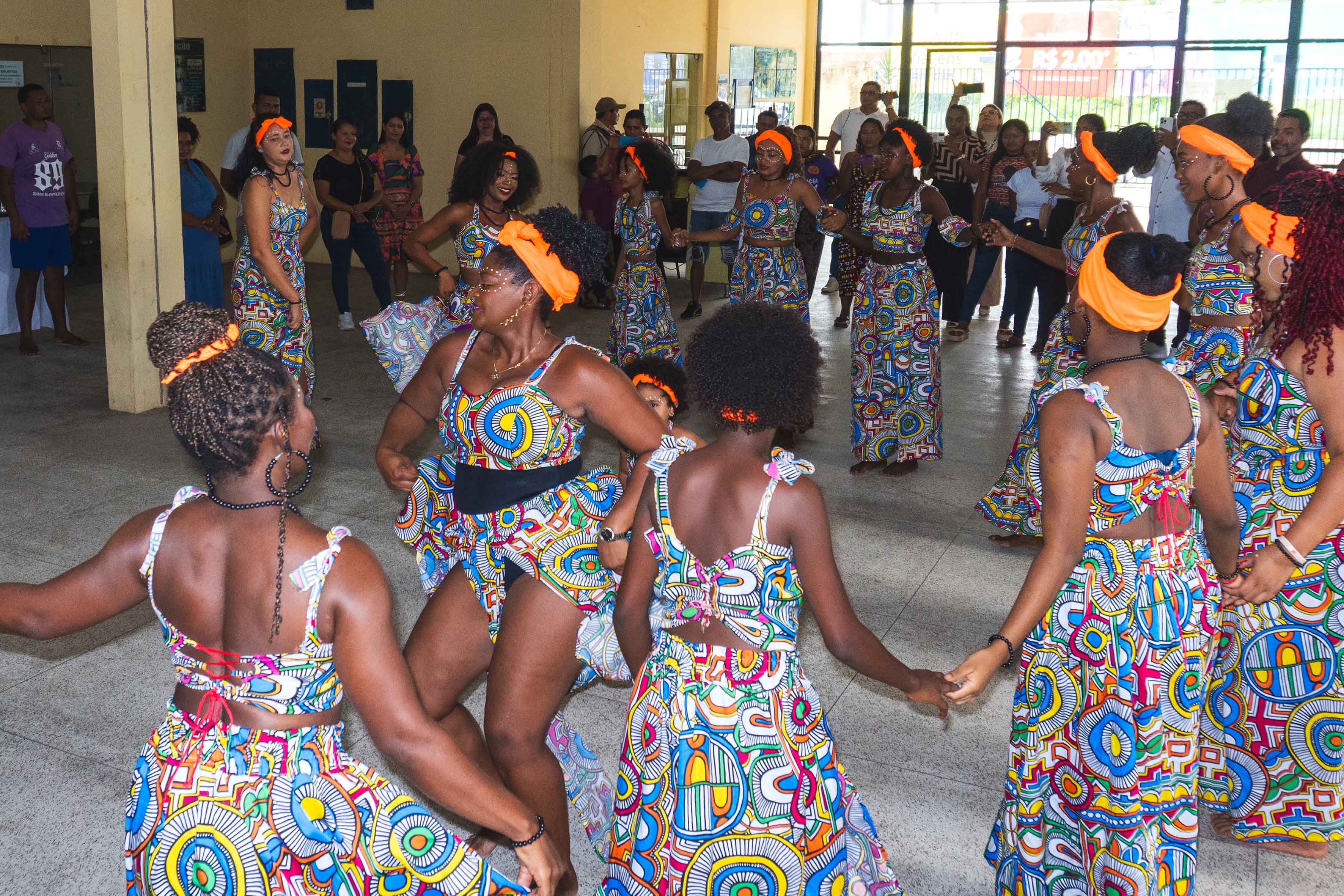 Na imagem, um grupo de mulheres negras dança em roda dentro de um espaço amplo e iluminado. Elas vestem roupas coloridas com estampas afro e usam adornos como faixas laranja na cabeça e acessórios artesanais. A cena destaca a força da cultura afro-brasileira e o espírito coletivo da atividade.