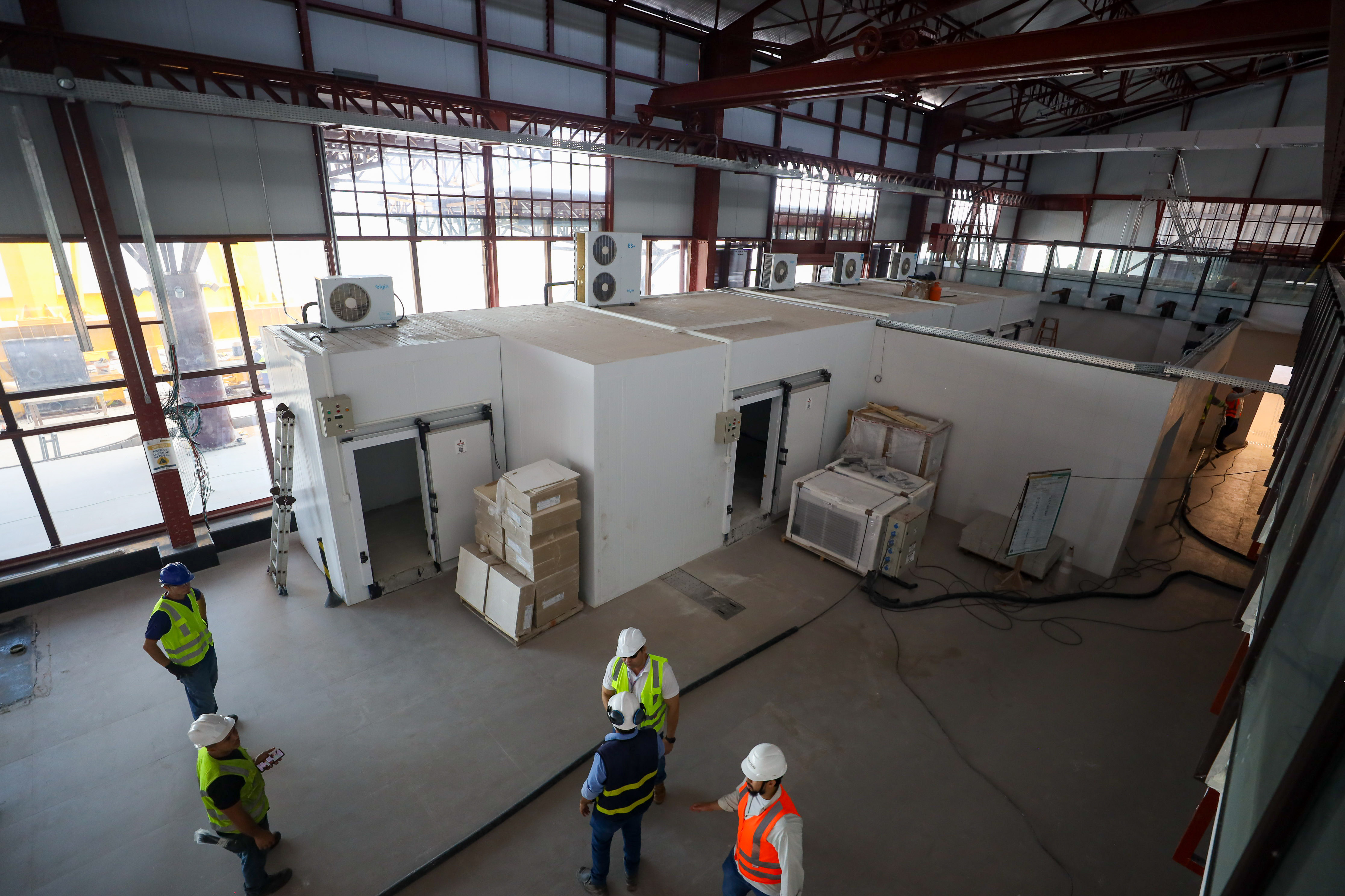 Vista interna de uma estrutura em fase de instalação ou adequação, com módulos de salas climatizadas de paredes brancas dispostos no centro do ambiente. No local, trabalhadores utilizam equipamentos de proteção individual enquanto acompanham as obras. A área possui pé-direito elevado, estrutura metálica aparente, grandes painéis de vidro e equipamentos de climatização instalados sobre os módulos.