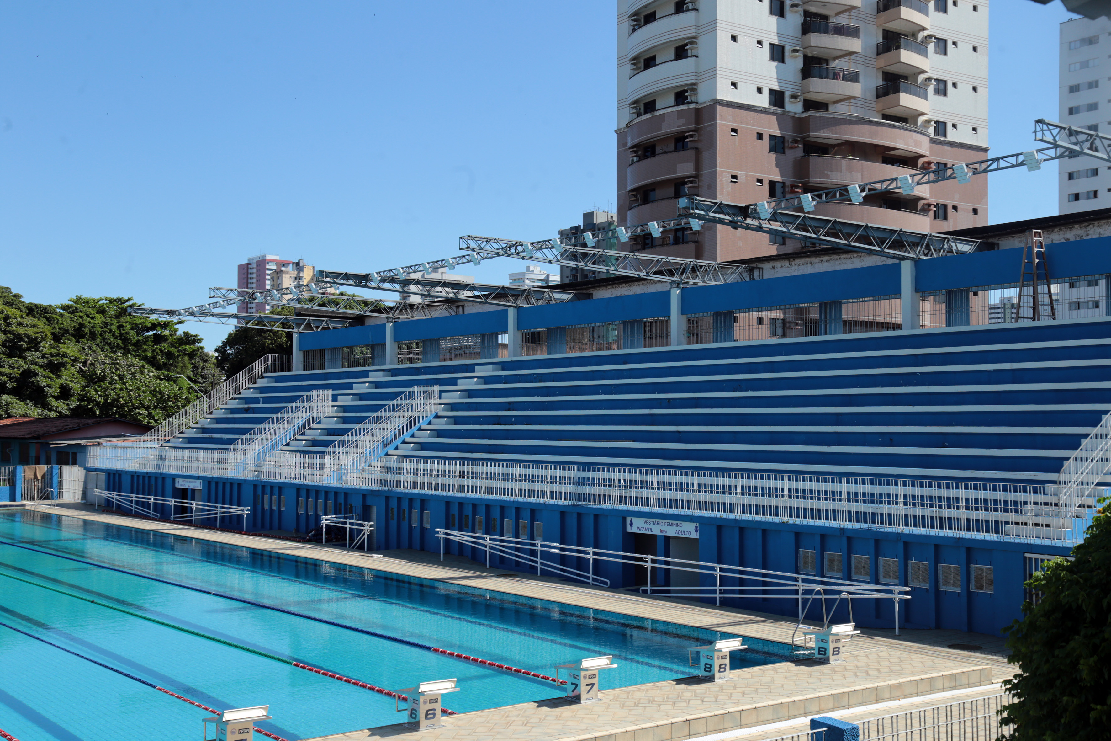 Piscina ao ar livre com raias para competição. Ao redor da piscina, há arquibancadas azuis para os espectadores. Acima das arquibancadas,  há uma estrutura em reforma. Ao fundo, céu claro.