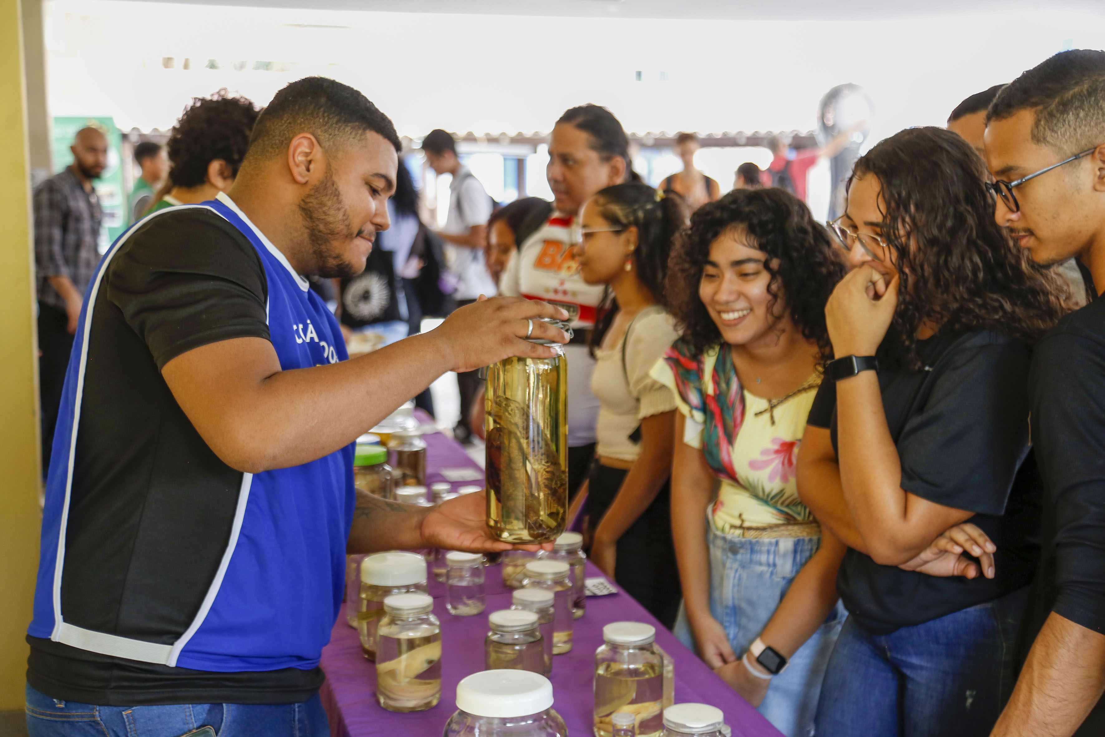  A imagem mostra um grupo de pessoas em um ambiente de exposição ou feira. Um jovem, vestido com uma camiseta preta e um colete azul, está segurando um frasco grande com líquido e um objeto dentro, possivelmente um animal conservado em formol. Entre os espectadores, uma jovem com cabelo cacheado e outra com uma blusa estampada sorriem, demonstrando curiosidade. A mesa à frente está coberta com toalhas roxas e cheia de frascos menores, sugerindo uma apresentação sobre biologia ou conservação ambiental.