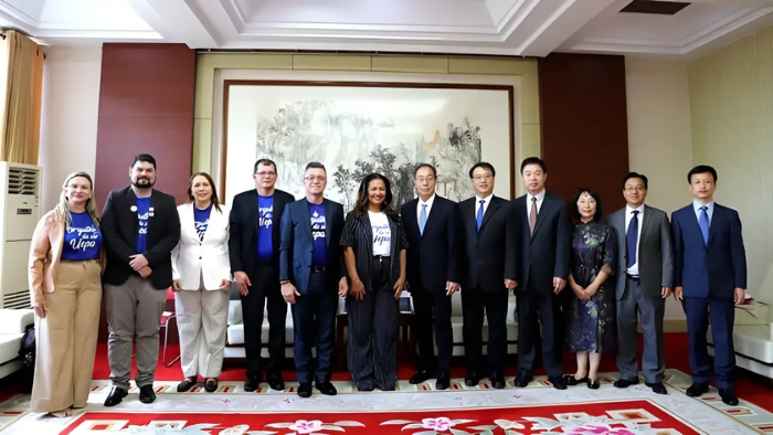 A imagem mostra um grupo de 11 pessoas posando para uma foto oficial em um ambiente interno. O cenário é elegante, com paredes em tom claro, detalhes em madeira e um grande quadro de paisagem ao fundo. No chão, há um tapete decorado em vermelho e branco. A composição transmite a ideia de um encontro institucional entre duas delegações.