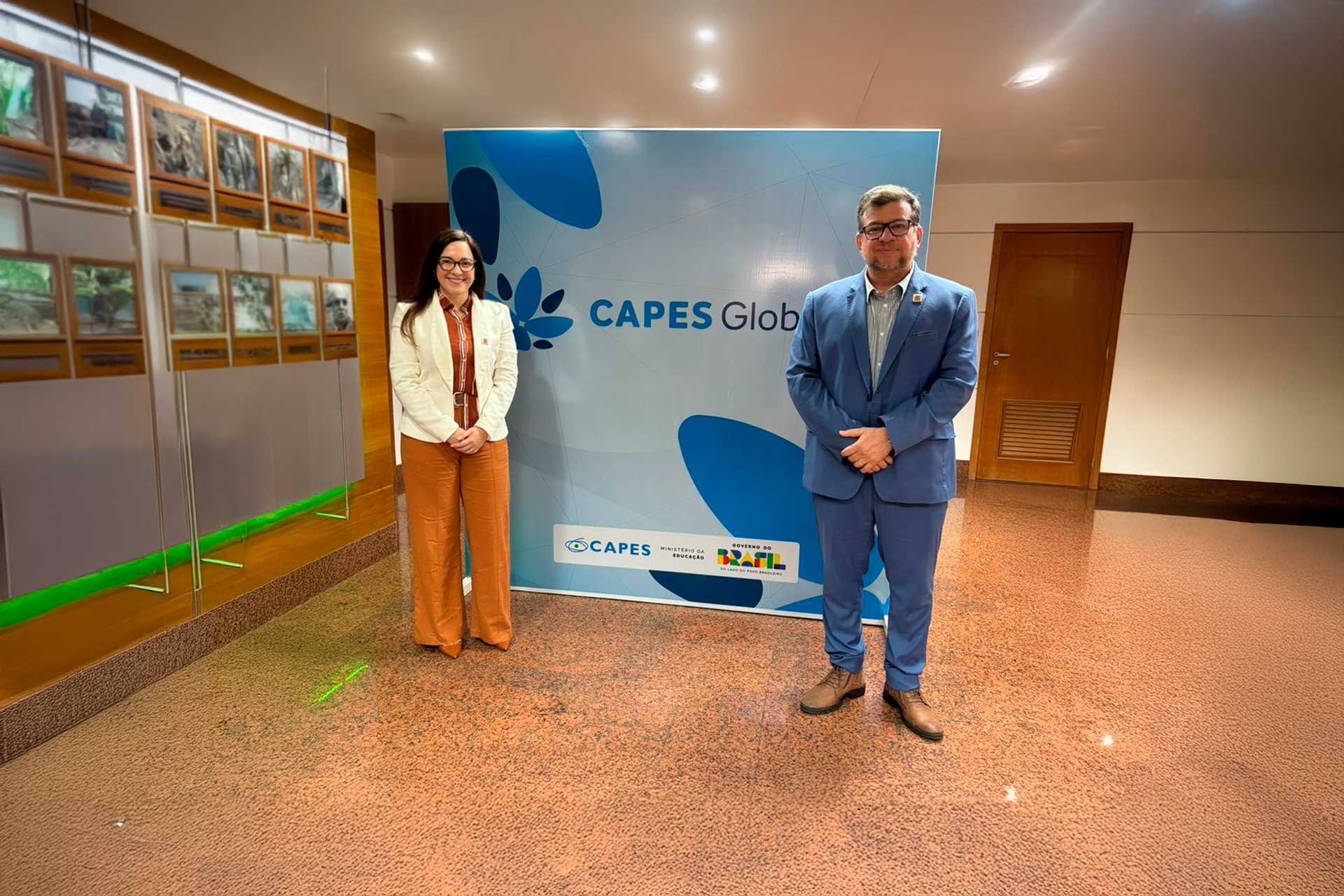 Duas pessoas posam em pé diante de um painel institucional com a marca “Capes Global”. Ambas vestem trajes formais e usam crachás, sugerindo participação em evento ou programa acadêmico. O ambiente é interno, com iluminação de escritório, piso em tom alaranjado e portas ao fundo, reforçando o caráter institucional da foto.