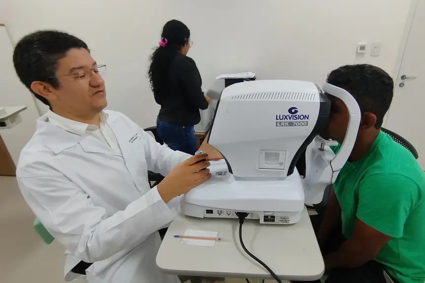 A imagem retrata um atendimento médico especializado em um ambiente clínico iluminado, onde um profissional de saúde, utilizando jaleco branco, realiza um exame oftalmológico em um paciente vestido com uma camiseta verde. O médico opera um equipamento de diagnóstico avançado,  ajustando o aparelho enquanto o paciente posiciona o rosto no suporte para a avaliação.