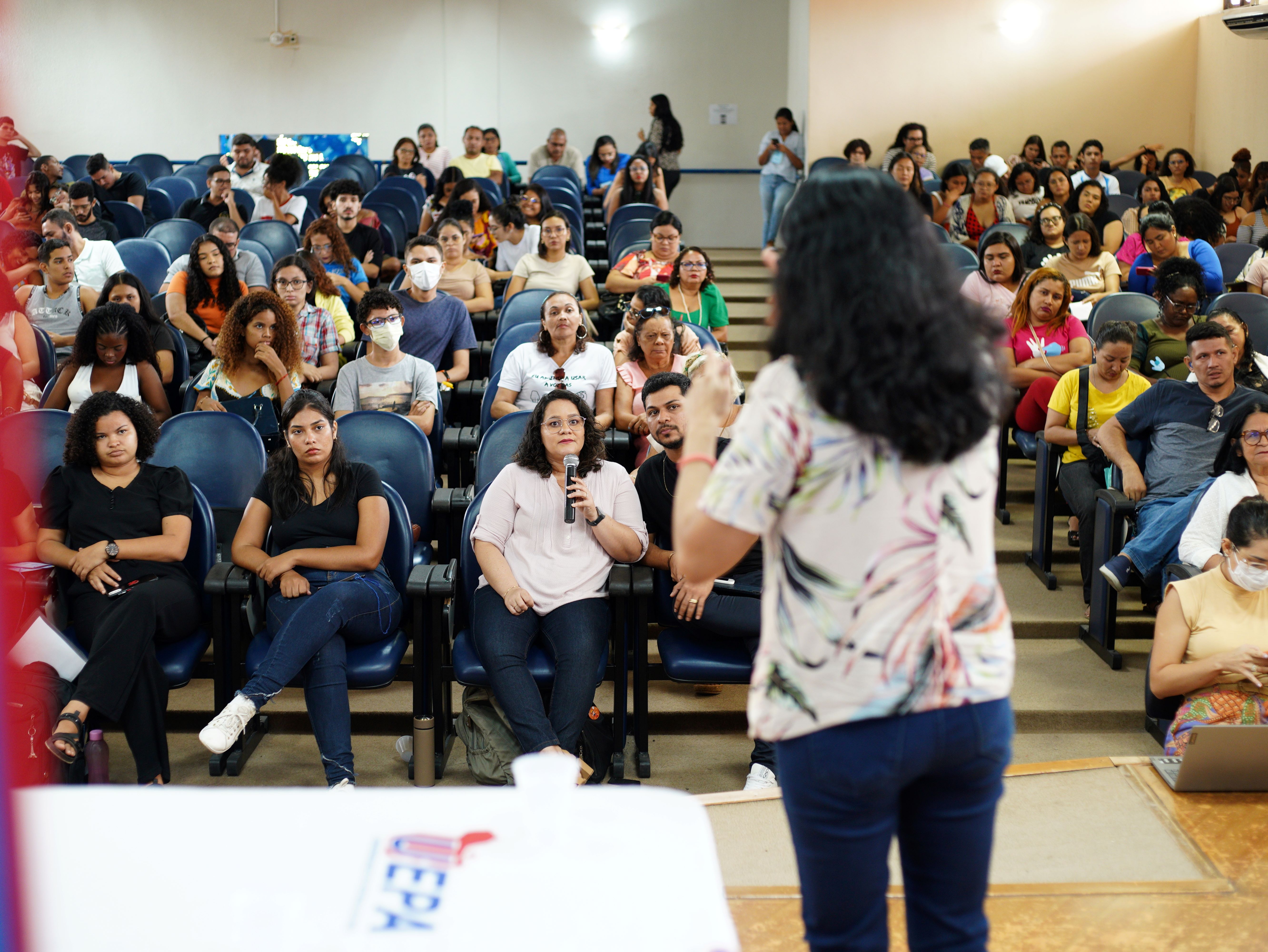 A imagem mostra um ambiente interno onde uma pessoa com cabelo comprido está de pé em frente a um grupo de indivíduos sentados, de frente para eles. A pessoa em primeiro plano parece estar se dirigindo ao público. O público é composto por pessoas de diferentes idades, sentadas em filas que se elevam em direção ao fundo da sala, o que sugere uma disposição em estilo de auditório. 