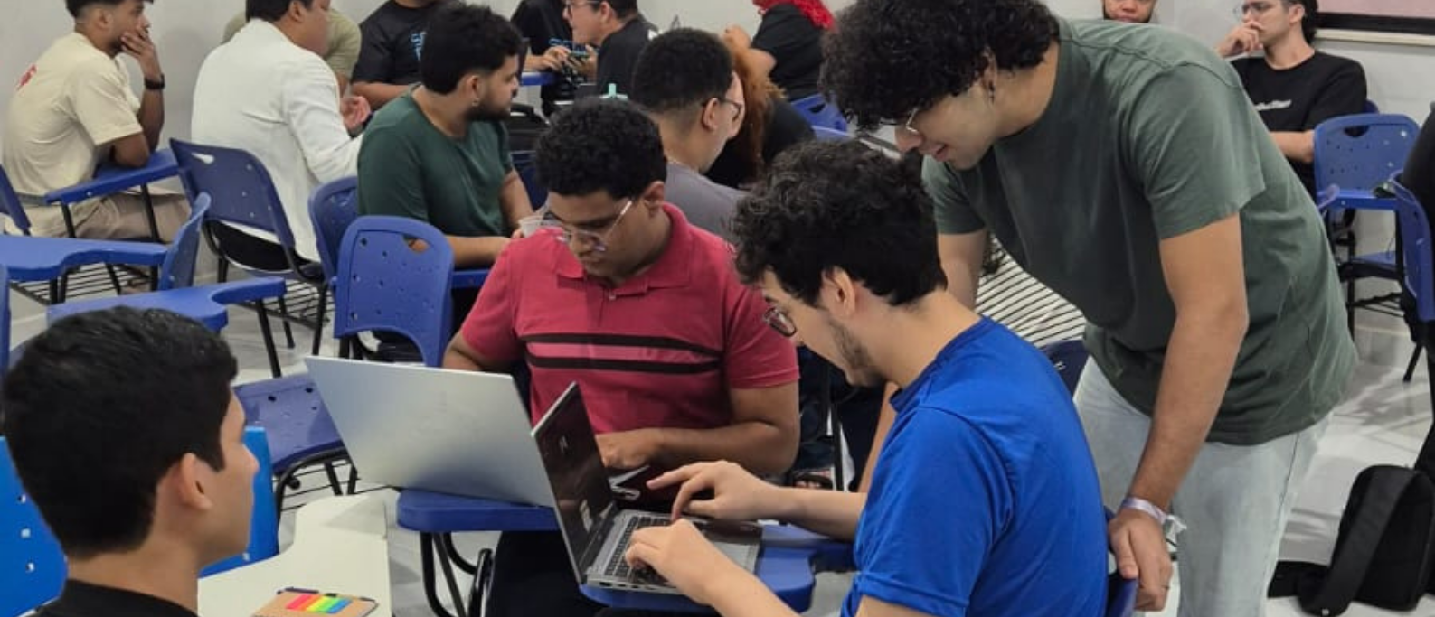 A imagem mostra uma sala de aula com grupos de estudantes reunidos em atividades colaborativas. As carteiras estão organizadas de forma a facilitar a interação, e vários alunos utilizam notebooks para desenvolver seus trabalhos.