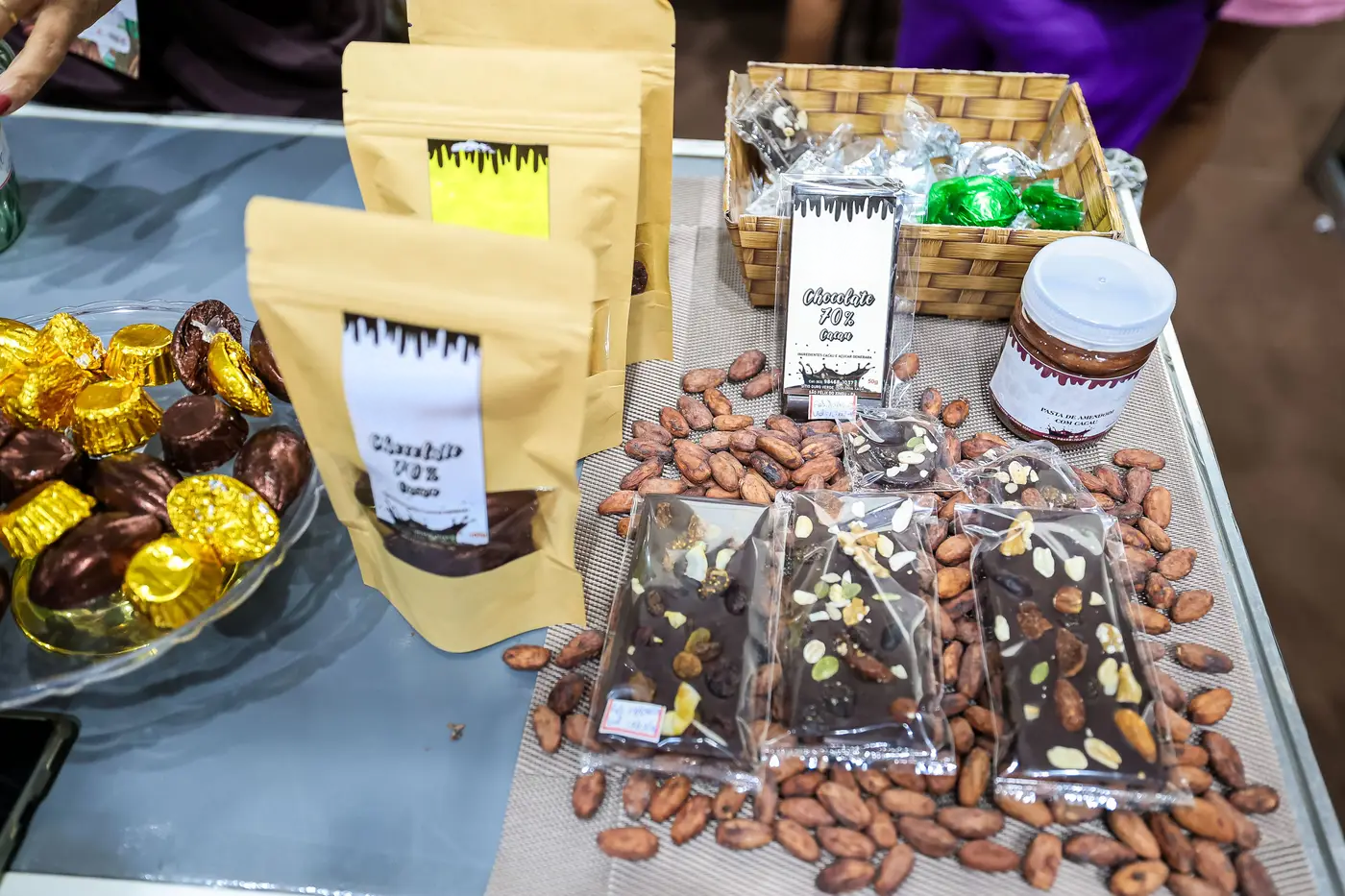Bancada de exposição em uma feira com diversos produtos de chocolate: barras artesanais embaladas, bombons dourados, amêndoas de cacau espalhadas, potes e uma cesta com itens embalados, destacando a apresentação de chocolates de diferentes tipos e intensidades.