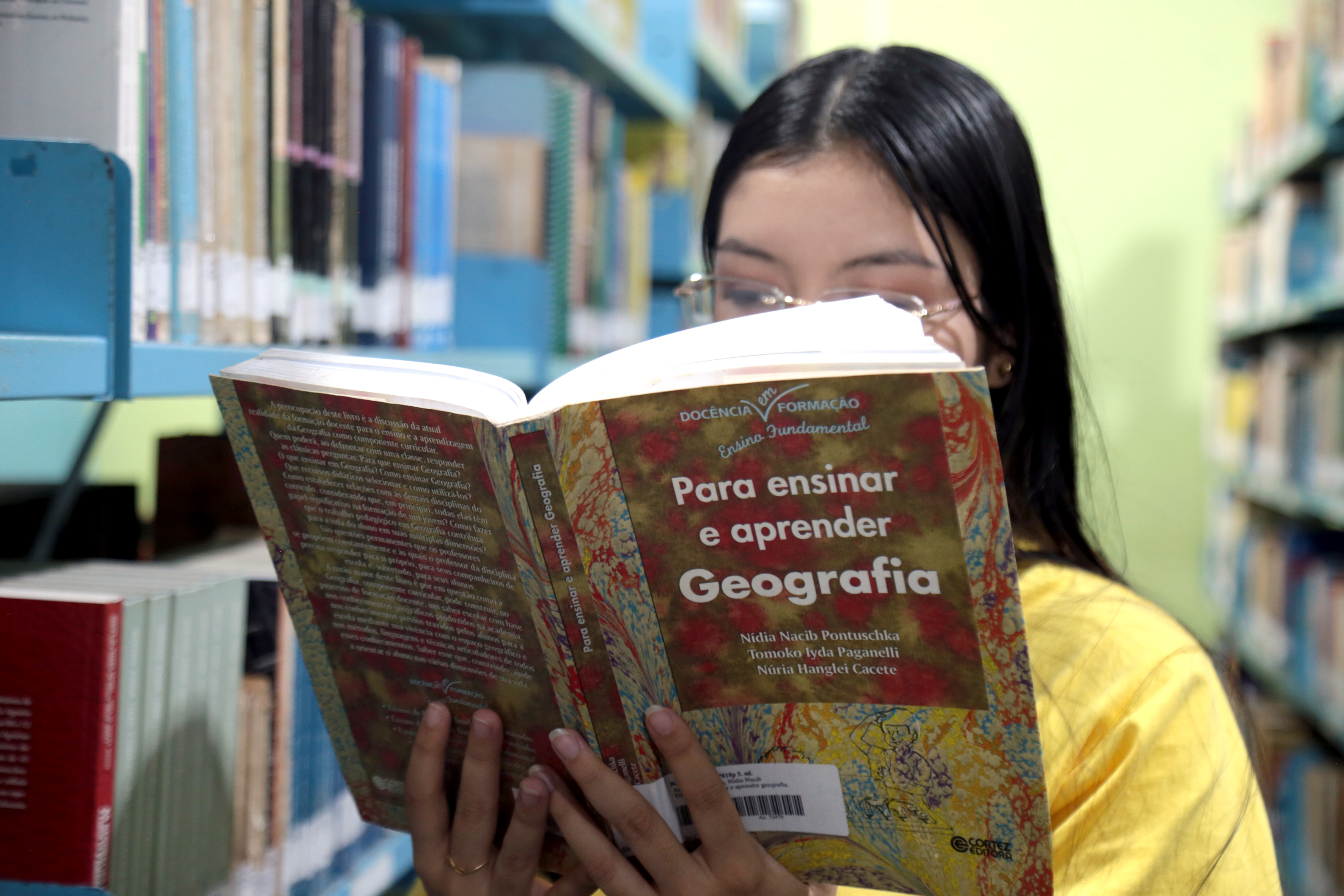 A imagem mostra uma pessoa em frente a uma estante de livros, lendo um livro intitulado “Geografia.”  A capa do livro apresenta várias cores e parece ter um mapa. Atrás da pessoa, há várias prateleiras cheias de livros, indicando que o local é uma biblioteca.