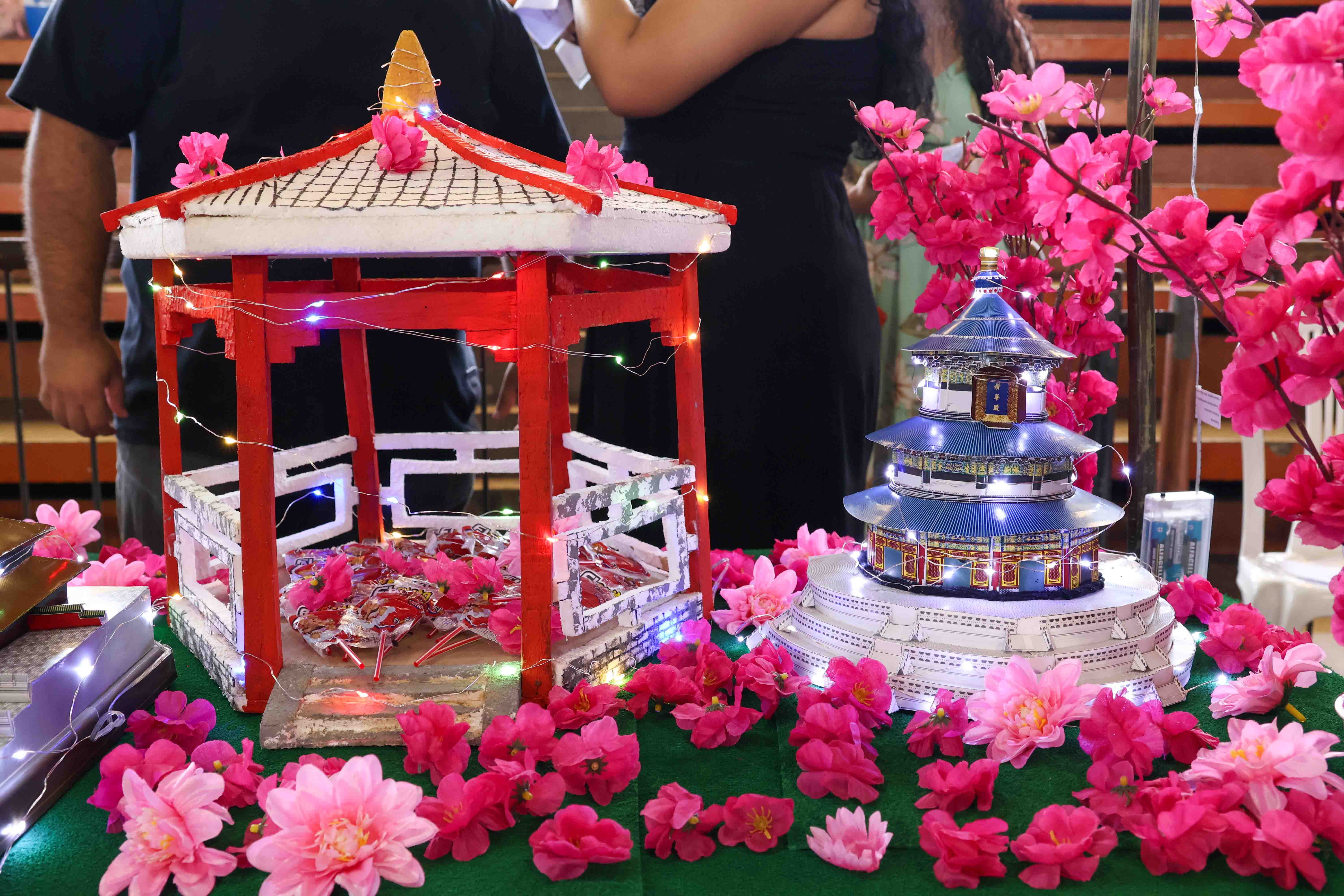 A imagem mostra uma maquete com elementos da cultura oriental. À esquerda, há uma estrutura com um telhado vermelho e branco, decorado com flores rosas artificiais e luzes coloridas. No centro, há pirulitos dispostos dentro dessa estrutura. À direita, um edifício tradicional asiático de vários andares, com telhados azuis e base branca, está iluminado. O cenário é cercado por flores de cerejeira artificiais, criando uma atmosfera festiva, com pessoas ao fundo observando a maquete.