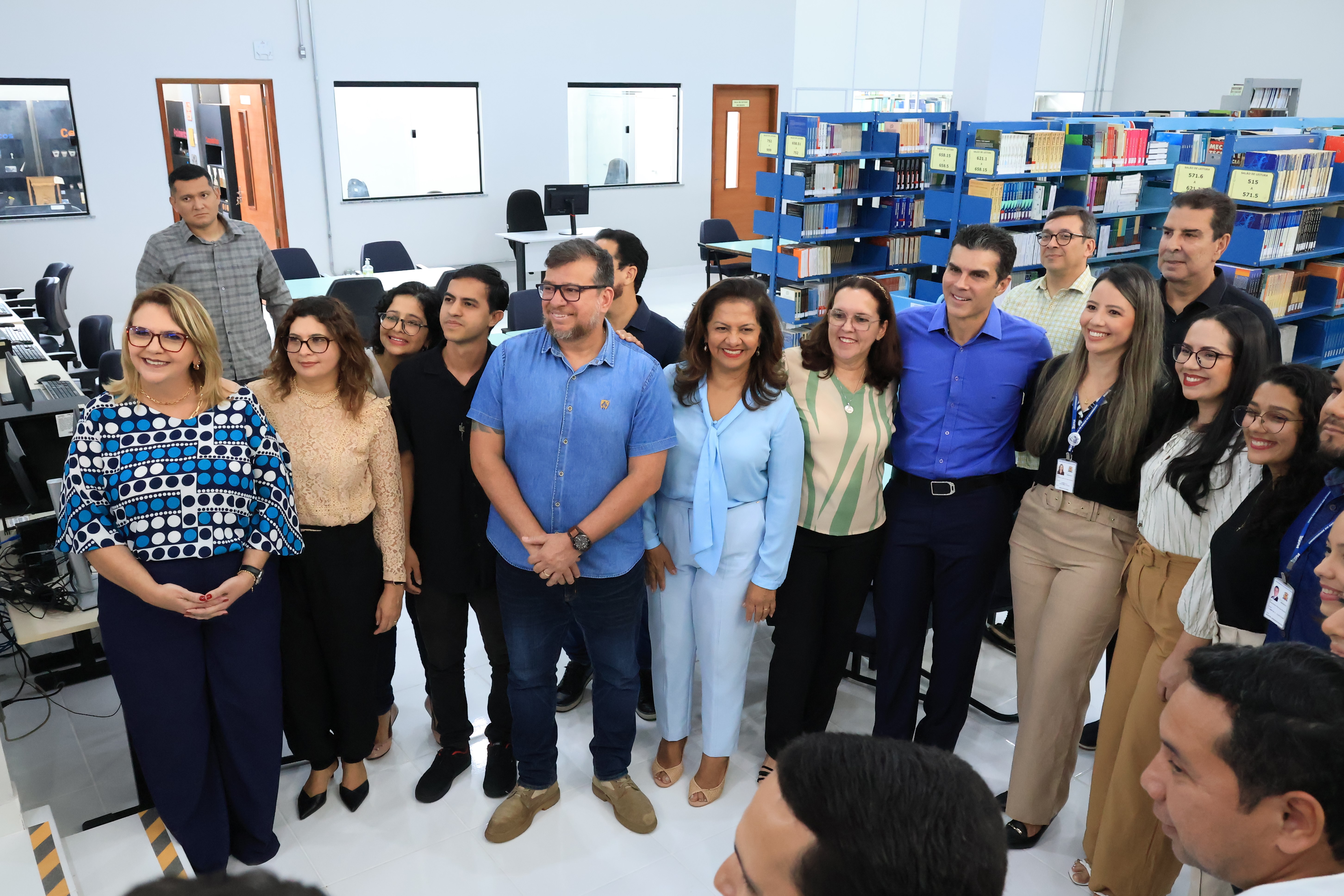 A imagem mostra um grupo de pessoas reunidas em um ambiente interno, aparentemente uma biblioteca ou espaço educacional. Ao fundo, há estantes organizadas com livros e computadores sobre mesas de trabalho. As pessoas, algumas vestindo crachás e roupas formais, sorriem para a câmera, demonstrando um momento de confraternização ou registro oficial. O local é iluminado e moderno, com um piso branco brilhante e janelas que permitem a entrada de luz natural.