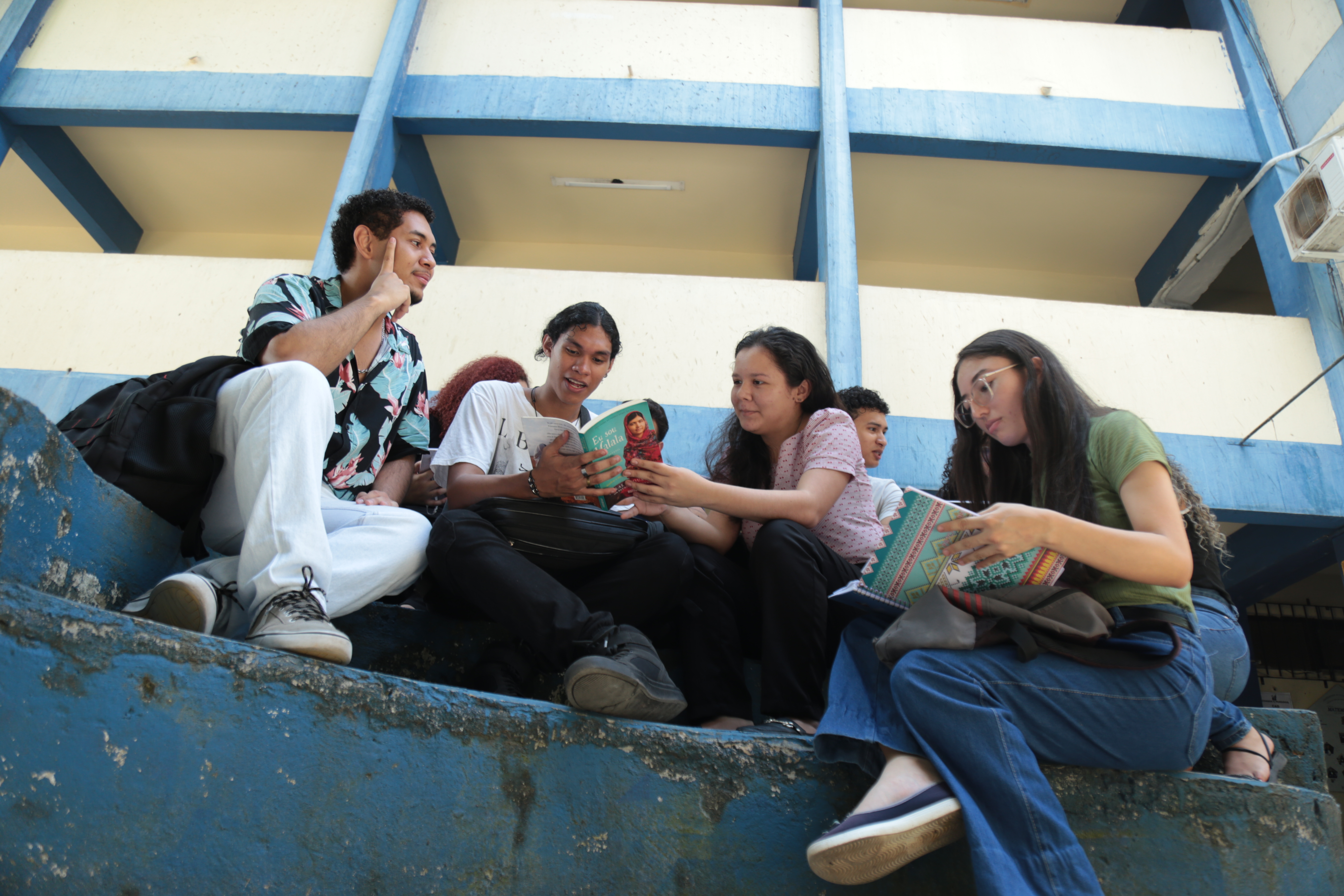 A imagem mostra um grupo de cinco pessoas sentadas em uma estrutura curva de concreto azul. Elas parecem estar envolvidas na leitura ou discussão de livros, já que várias delas estão segurando livros abertos. Ao fundo, há um prédio de vários andares com paredes azuis e amarelas. A cena sugere um ambiente casual, possivelmente educacional, onde os indivíduos são provavelmente estudantes ou amigos compartilhando um momento de estudo. Captura um momento de interação e aprendizado.