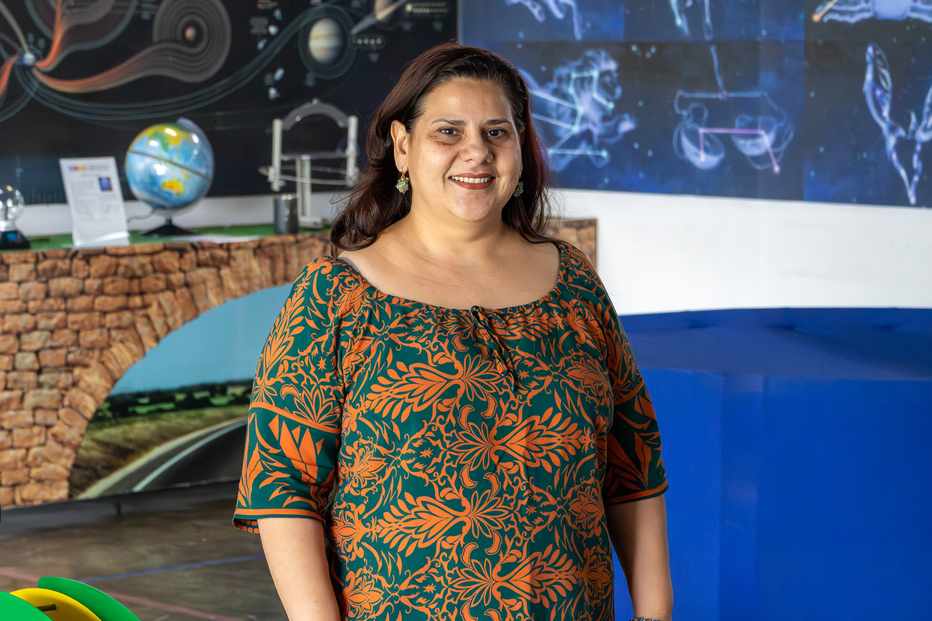 Professora sorri diante de um painel com ilustrações astronômicas. Ao fundo, há modelos científicos e um globo, compondo um ambiente educativo voltado para a ciência