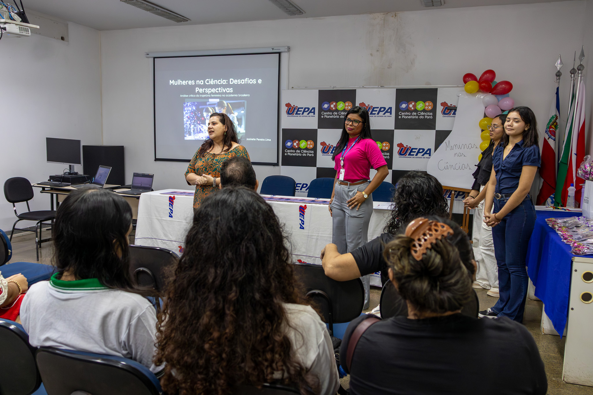 Três mulheres apresentam apresentam o projeto Meninas na Ciência, em sala decorada com banners institucionais. O público, sentado em cadeiras, acompanha atentamente a explicação.