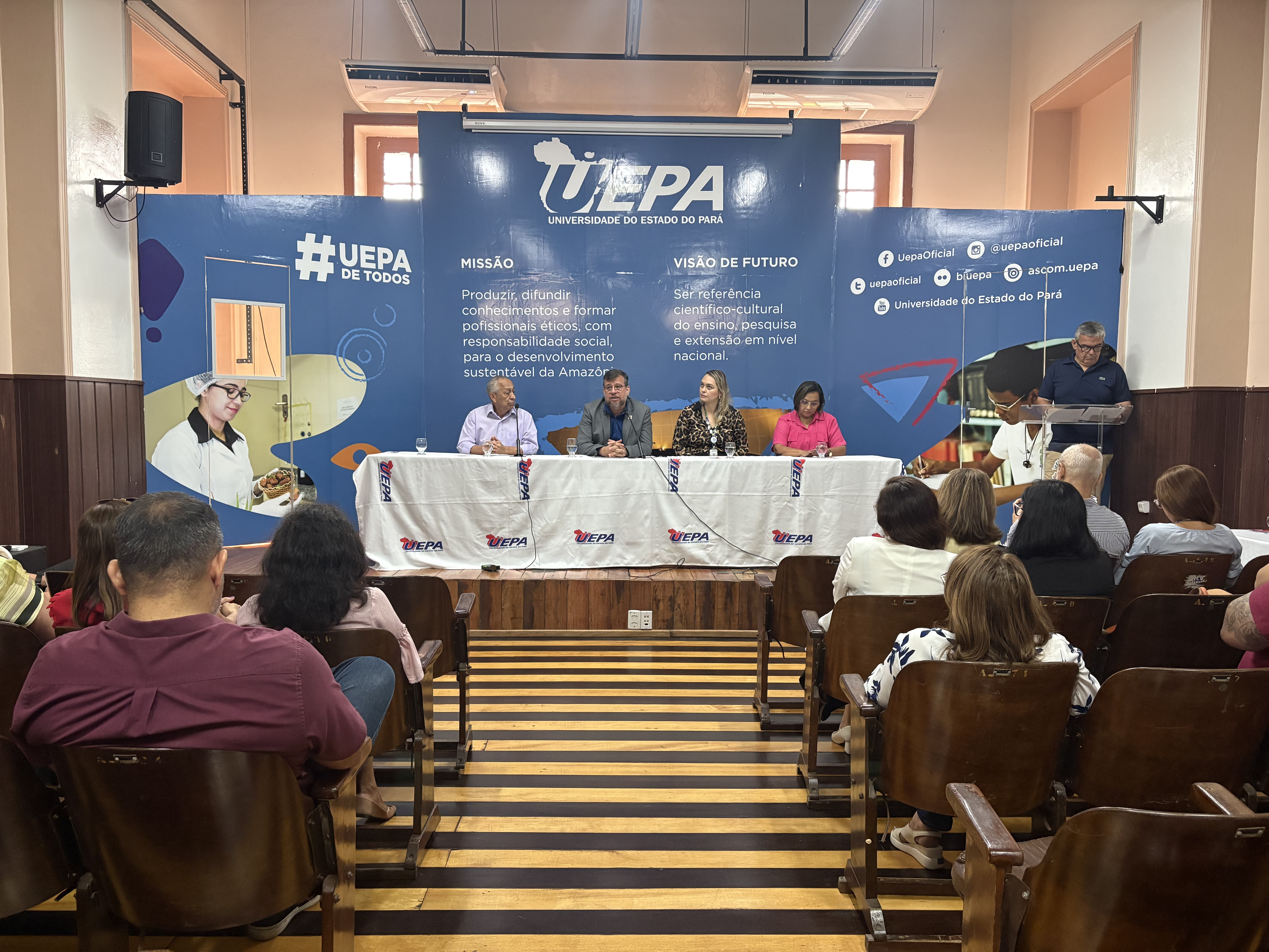 A imagem mostra um evento acadêmico com mesa composta e uma plateia atenta, em um ambiente formal.