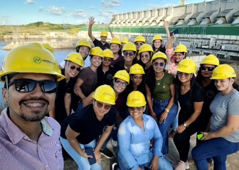 A imagem mostra um grupo de indivíduos usando capacetes de segurança amarelo, que estão em frente a uma grande  estrutura de uma represa, com portões de controle de água ao fundo, e retrata a turma do curso de Ciências Naturais após a visita técnica a Usina de Belo Monte.