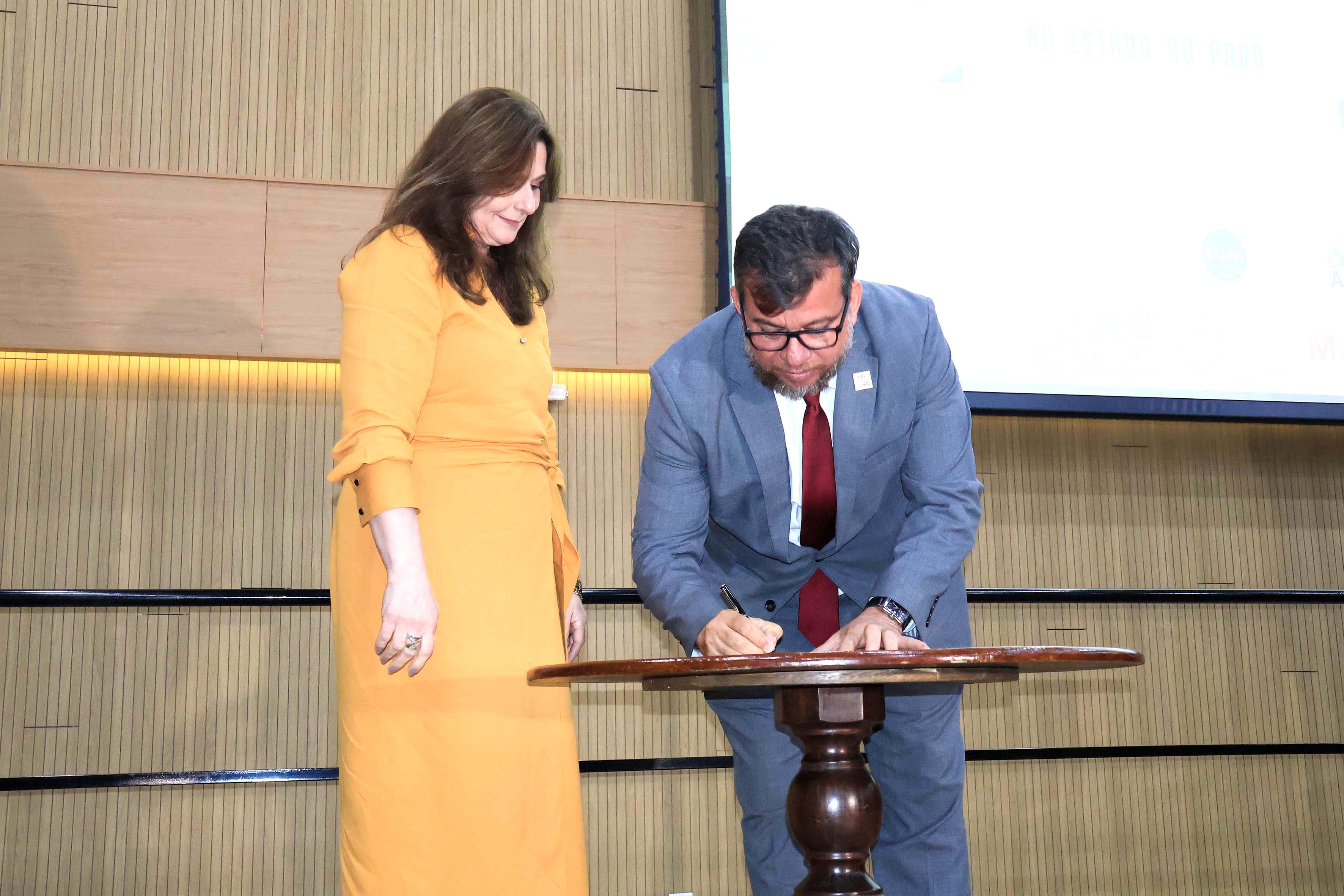 A imagem mostra duas pessoas em um ambiente formal, durante uma cerimônia de assinatura. À direita, um homem de terno cinza, gravata vermelha e óculos está assinando um documento em uma mesa pequena e redonda de madeira. Ao lado dele, uma mulher de vestido longo amarelo observa a assinatura, sorrindo levemente. O fundo apresenta uma parede de madeira clara com uma iluminação suave, indicando um local interno, possivelmente uma sala de conferências ou auditório.