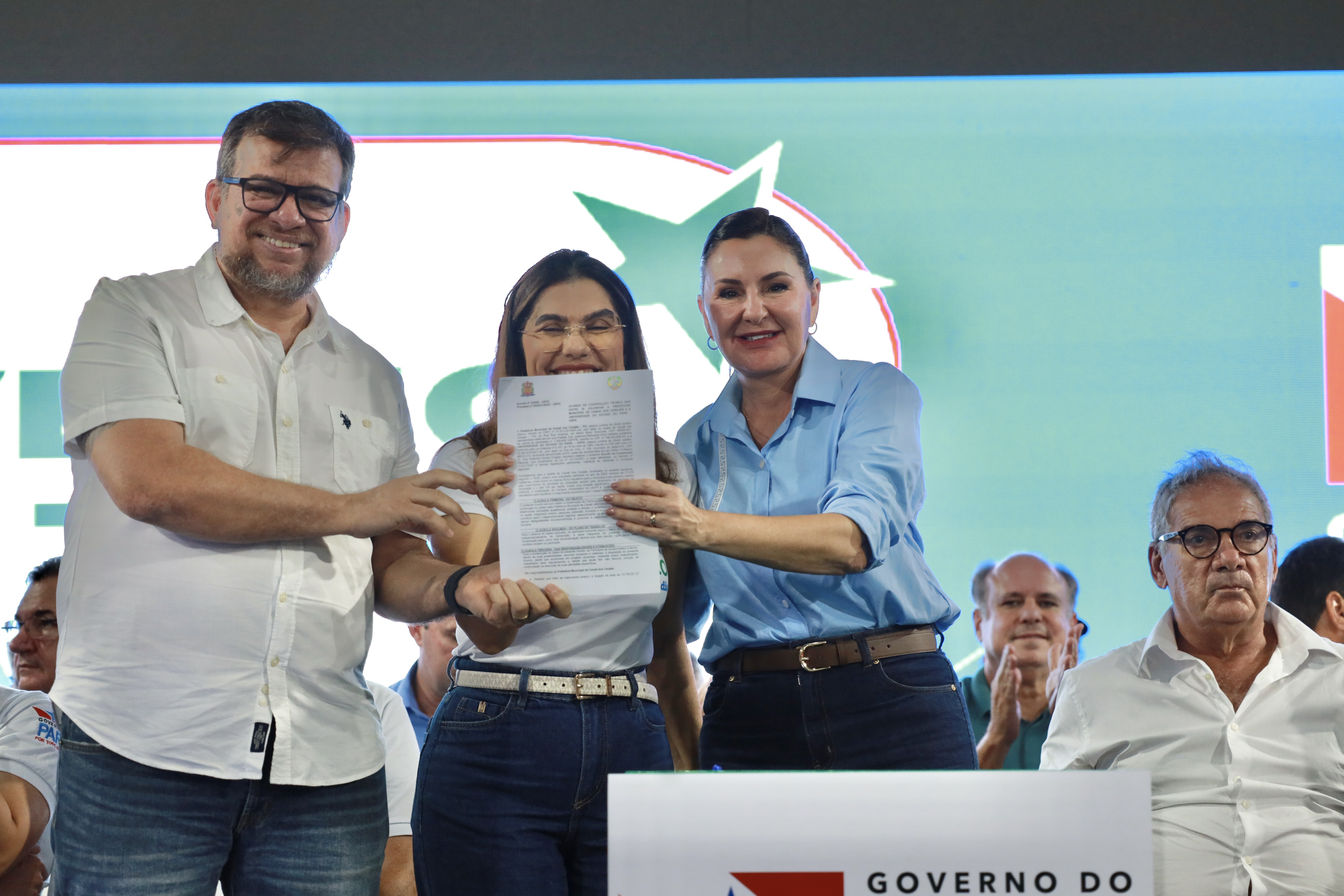 Três pessoas estão em destaque sobre um palco, sorrindo e segurando juntas um documento oficial voltado para a câmera. Ao fundo, há um painel iluminado com elementos gráficos e outras pessoas sentadas, algumas aplaudindo, indicando um evento institucional