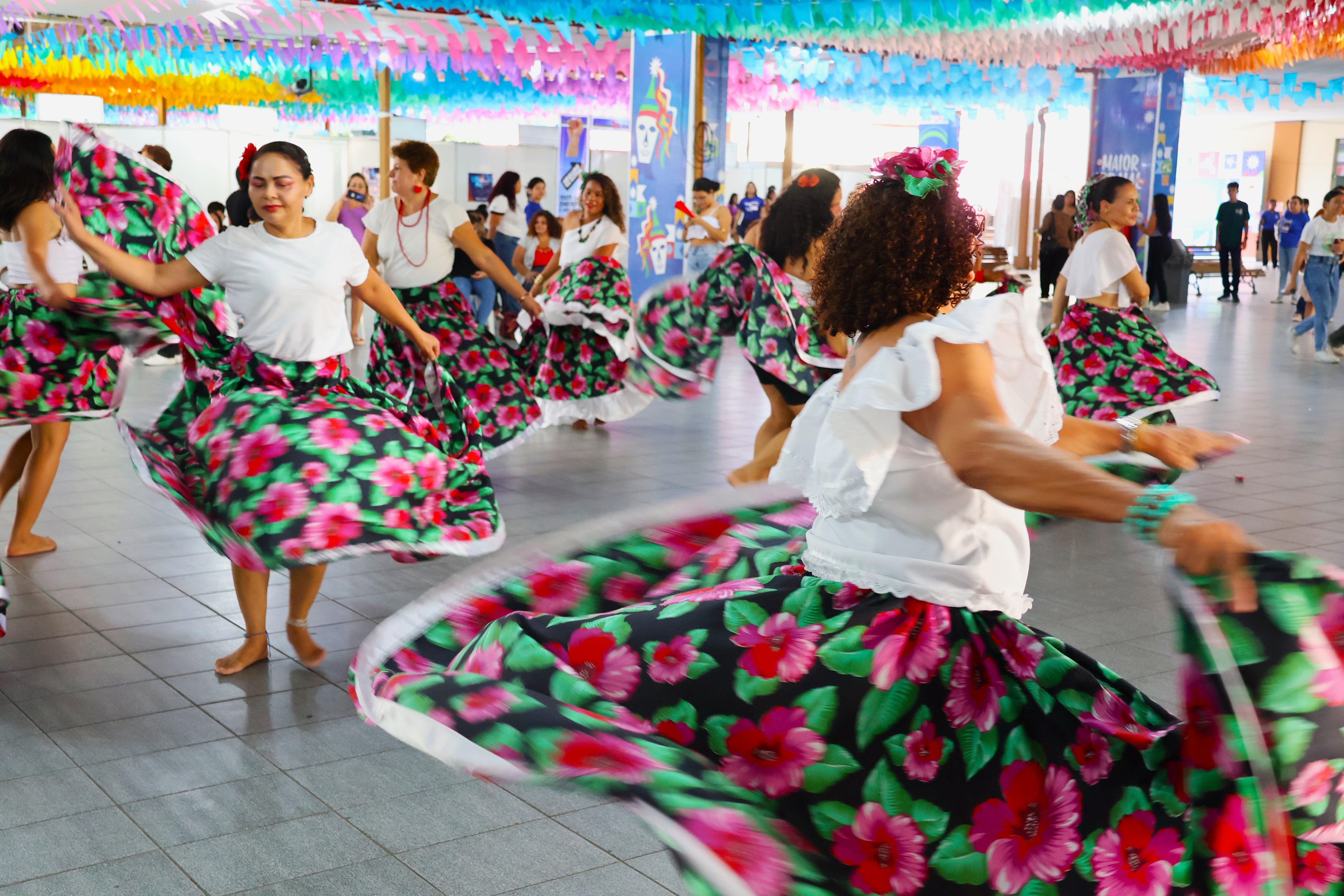 A imagem mostra um grupo de mulheres dançando em um ambiente decorado com bandeirinhas coloridas. Elas vestem saias rodadas estampadas com flores grandes e coloridas, combinadas com blusas brancas. O movimento das saias cria um efeito vibrante e dinâmico.