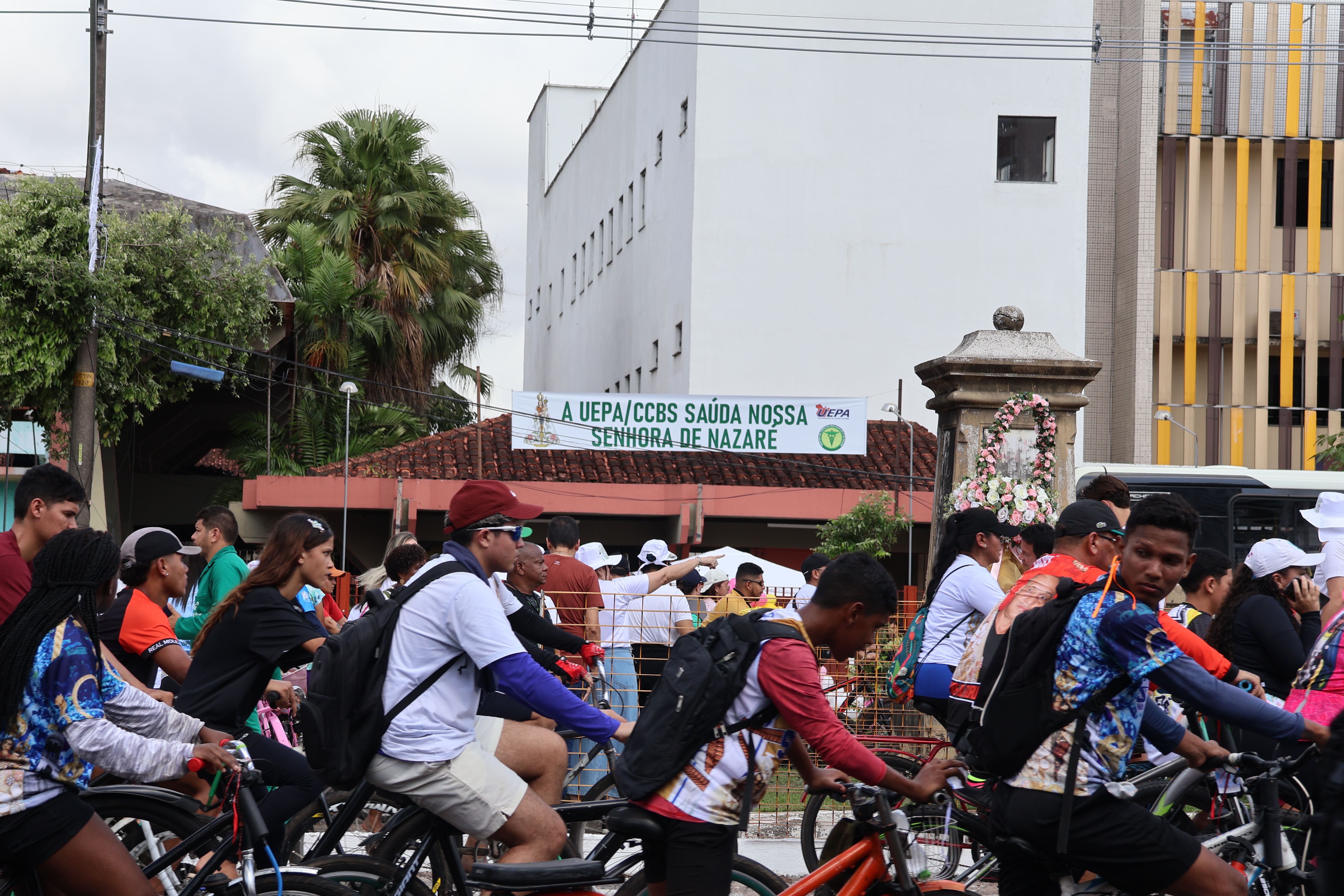A imagem mostra um grupo de ciclistas participando de uma romaria. Ao fundo, há uma faixa saudando Nossa Senhora de Nazaré, destacando a participação da Universidade no Círio de Nazaré.