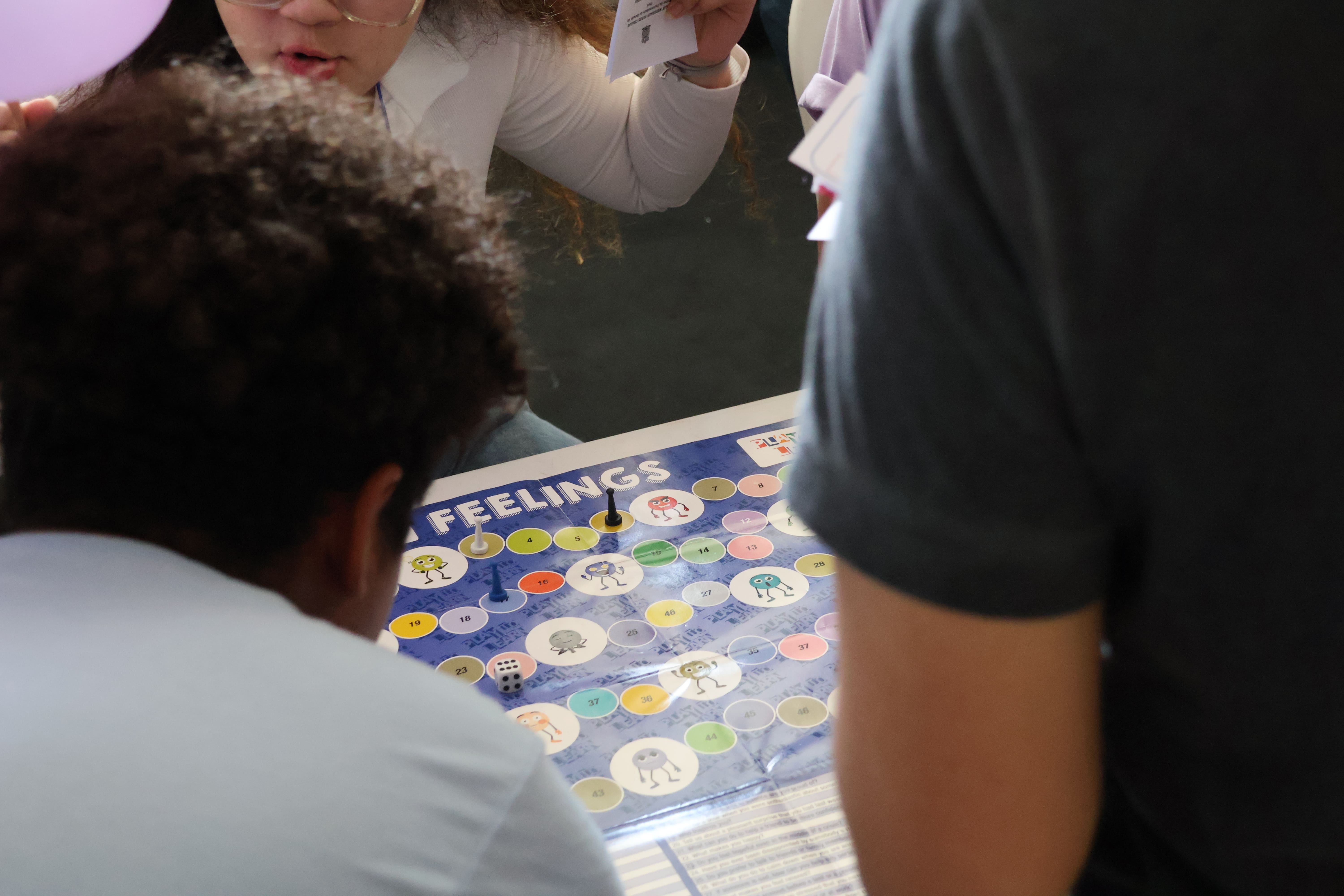 A imagem mostra três pessoas reunidas em torno de uma mesa, observando um jogo de tabuleiro chamado “Feelings”. O tabuleiro é colorido, com casas numeradas e personagens ilustrados que representam emoções. Há peões posicionados nas casas e um dado sobre o tabuleiro, indicando que a partida está em andamento. A cena sugere uma atividade interativa e educativa.