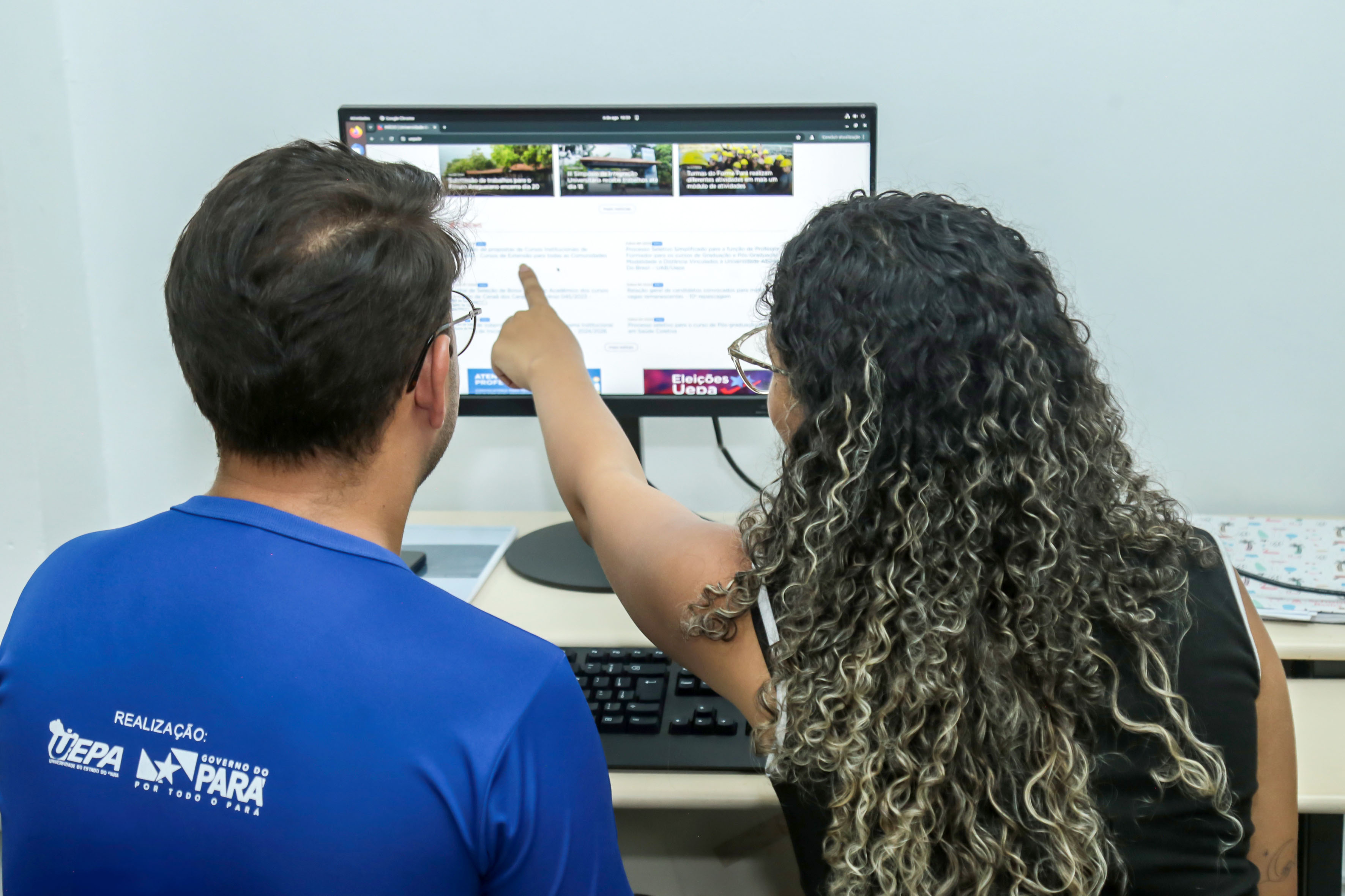 A imagem mostra duas pessoas em frente a um monitor de computador. Uma delas, com cabelos encaracolados e longos, está apontando para a tela do monitor, que exibe uma interface de usuário com vários elementos gráficos e textuais. A outra pessoa está olhando para a tela, acompanhando o que está sendo apontado. Ambas as pessoas estão vestindo camisetas azuis. O ambiente parece ser um escritório ou sala de aula com paredes brancas ao fundo.