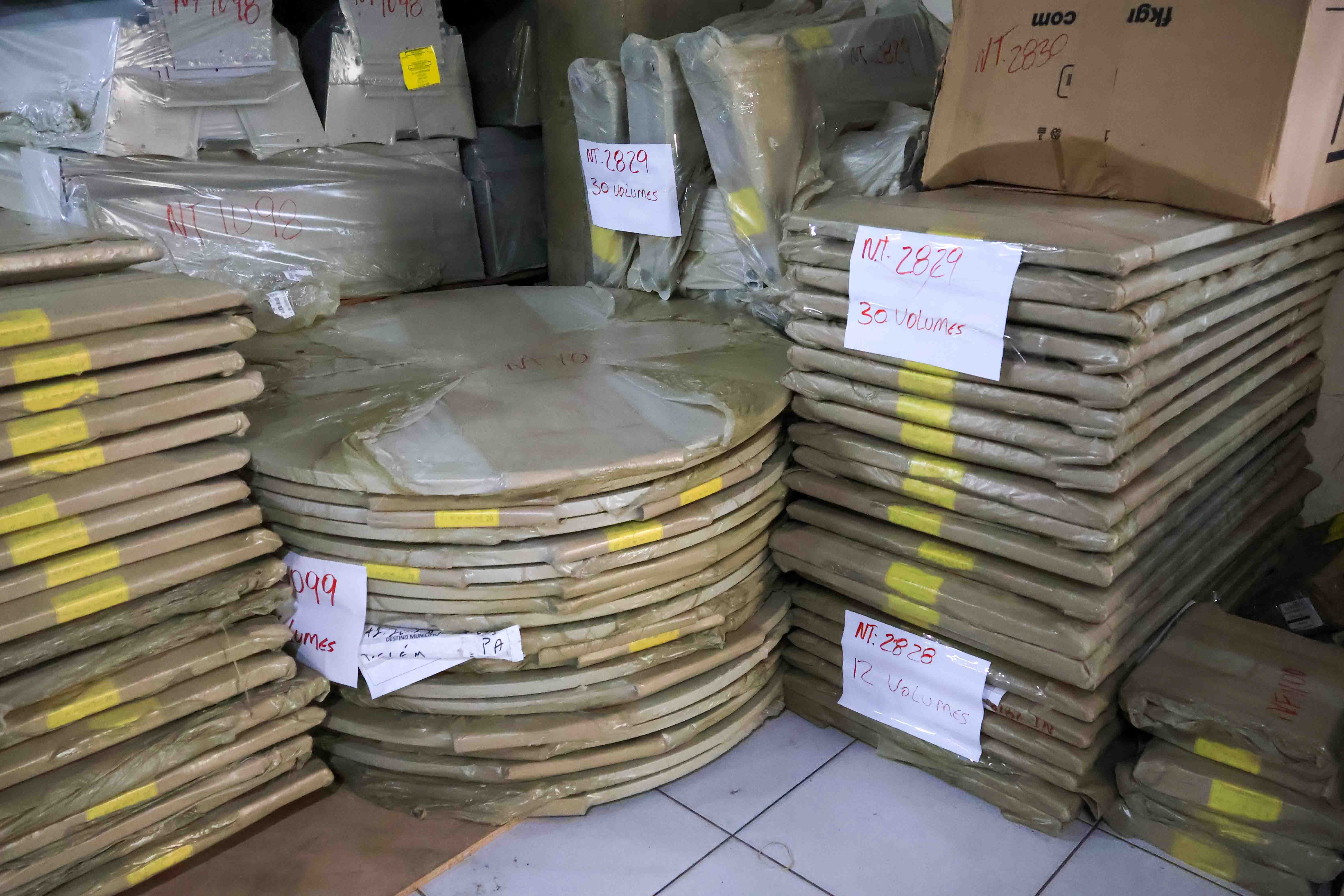 A imagem mostra pilhas de materiais embalados em papel pardo e plástico, organizados em um ambiente fechado. Algumas etiquetas identificam volumes e números de referência. Entre os itens, há mesas redondas e tampos retangulares, indicando possível mobiliário para bibliotecas ou espaços institucionais.